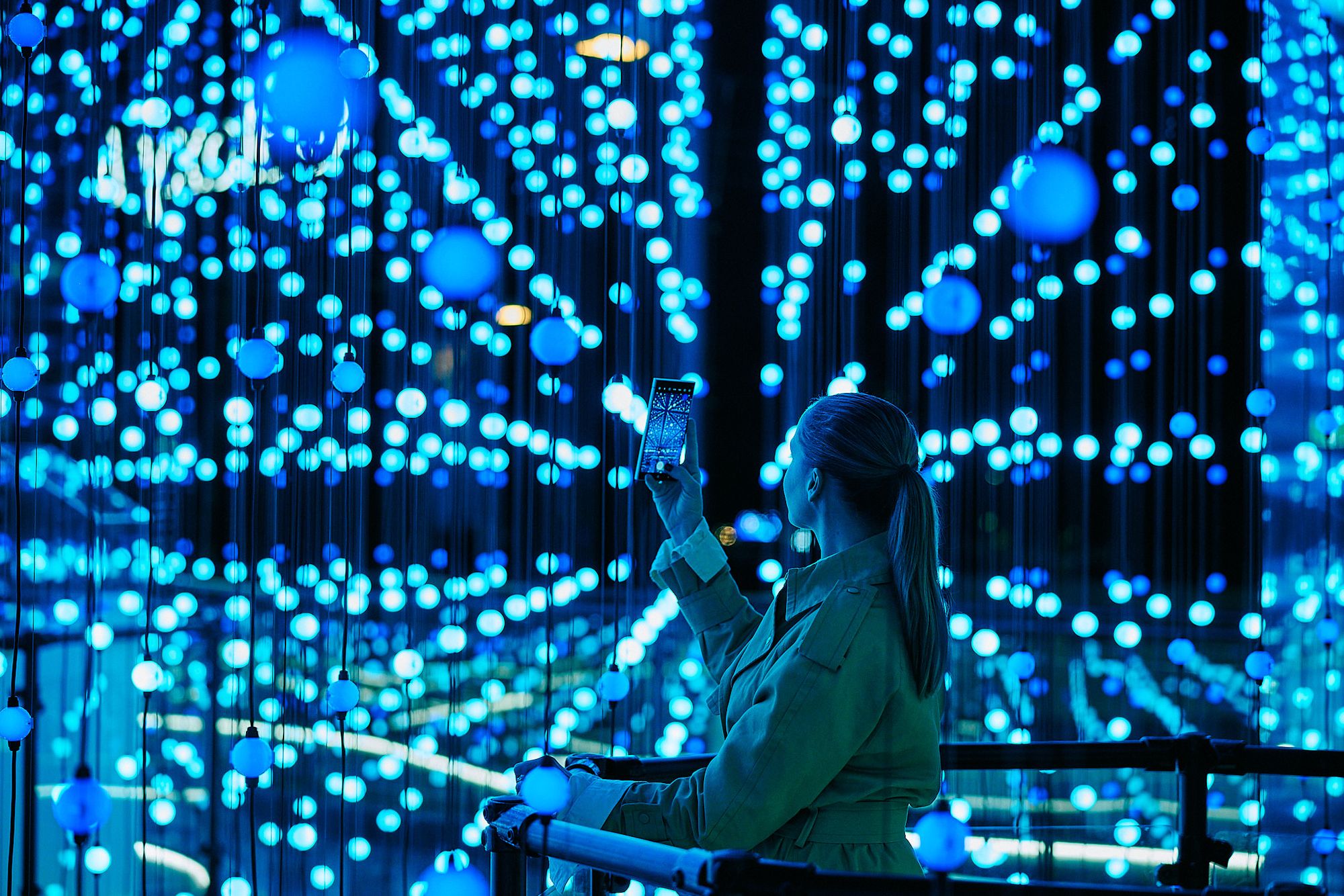 A woman stands in profile, silhouetted against an overwhelming galaxy of electric blue and cyan light. She holds a Samsung Galaxy smartphone raised above the railing in front of her, capturing the installation on her device — her screen glowing with the same brilliant light that surrounds her entirely.