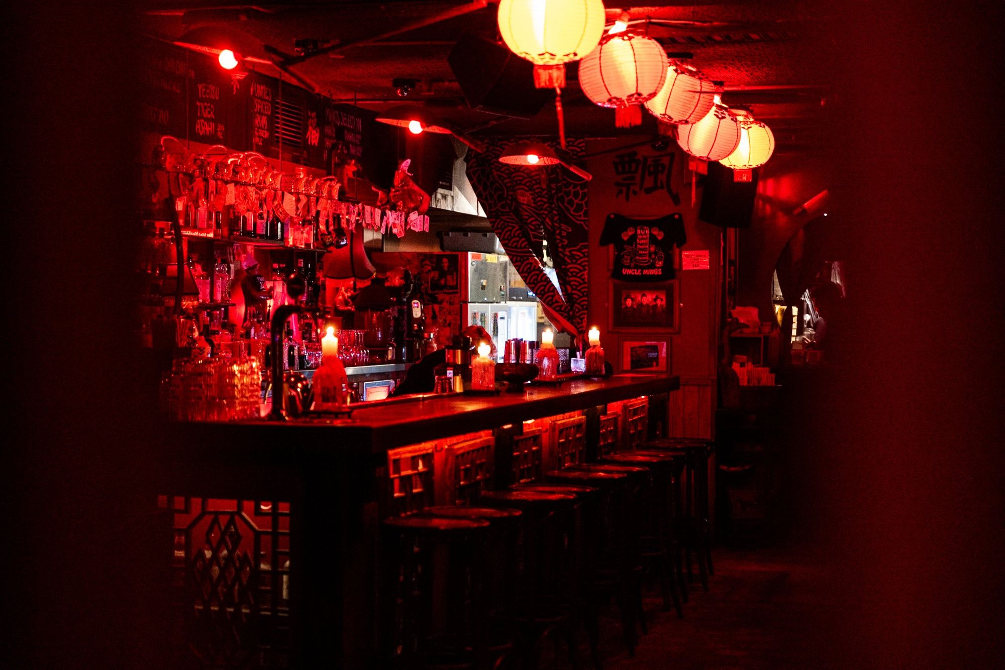 A dimly lit bar with red lanterns, wooden stools, and eclectic decorations creates an intimate atmosphere.