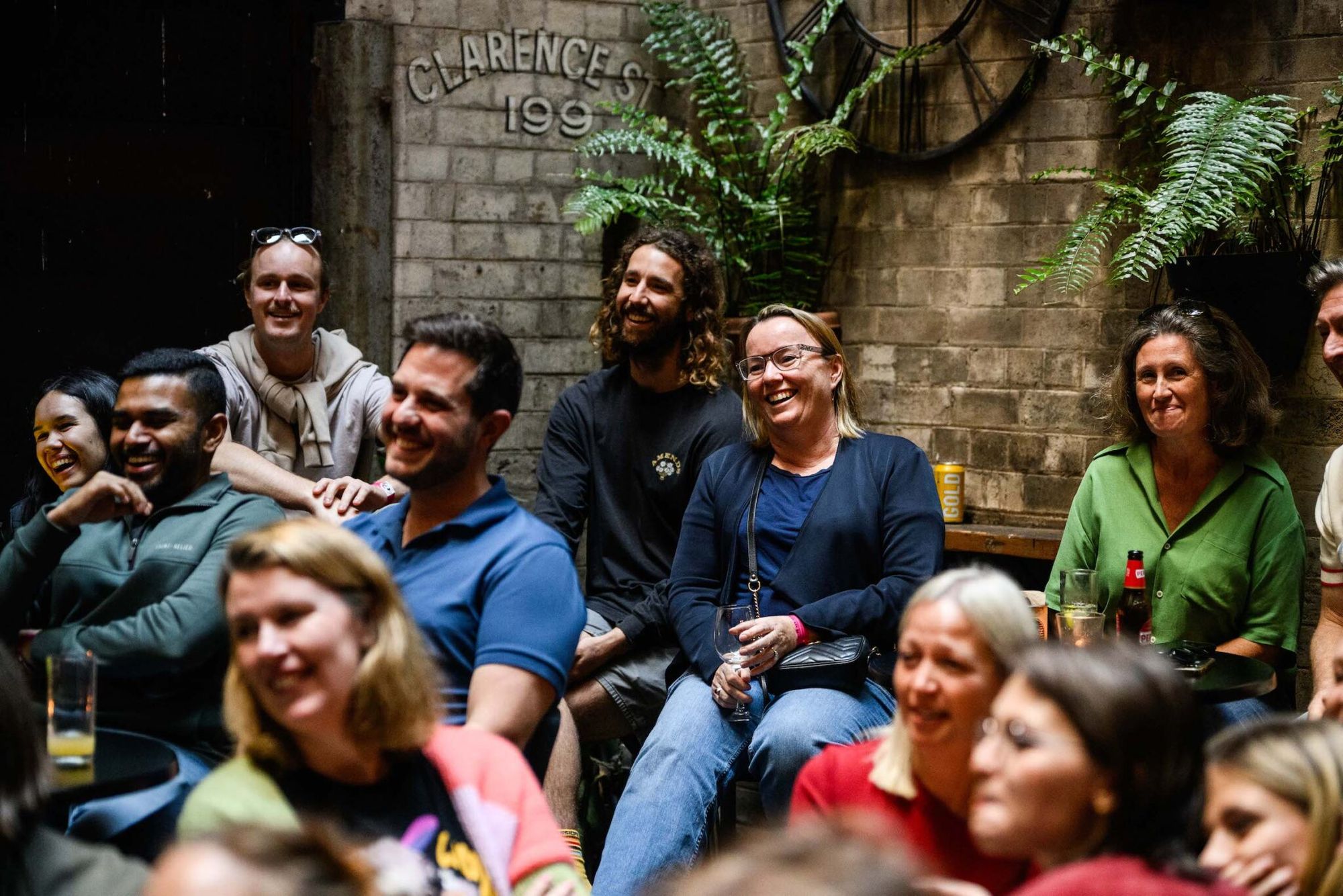 A group of people sitting together, smiling and laughing, in an indoor gathering with ferns and a brick wall in the background.