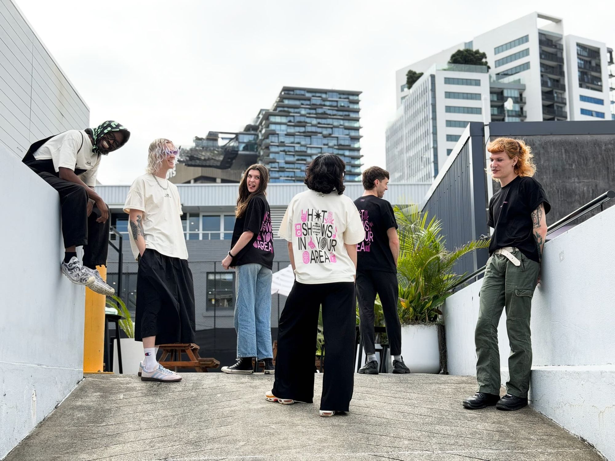 A group of six people in casual streetwear gather on an urban rooftop, with modern buildings in the background.