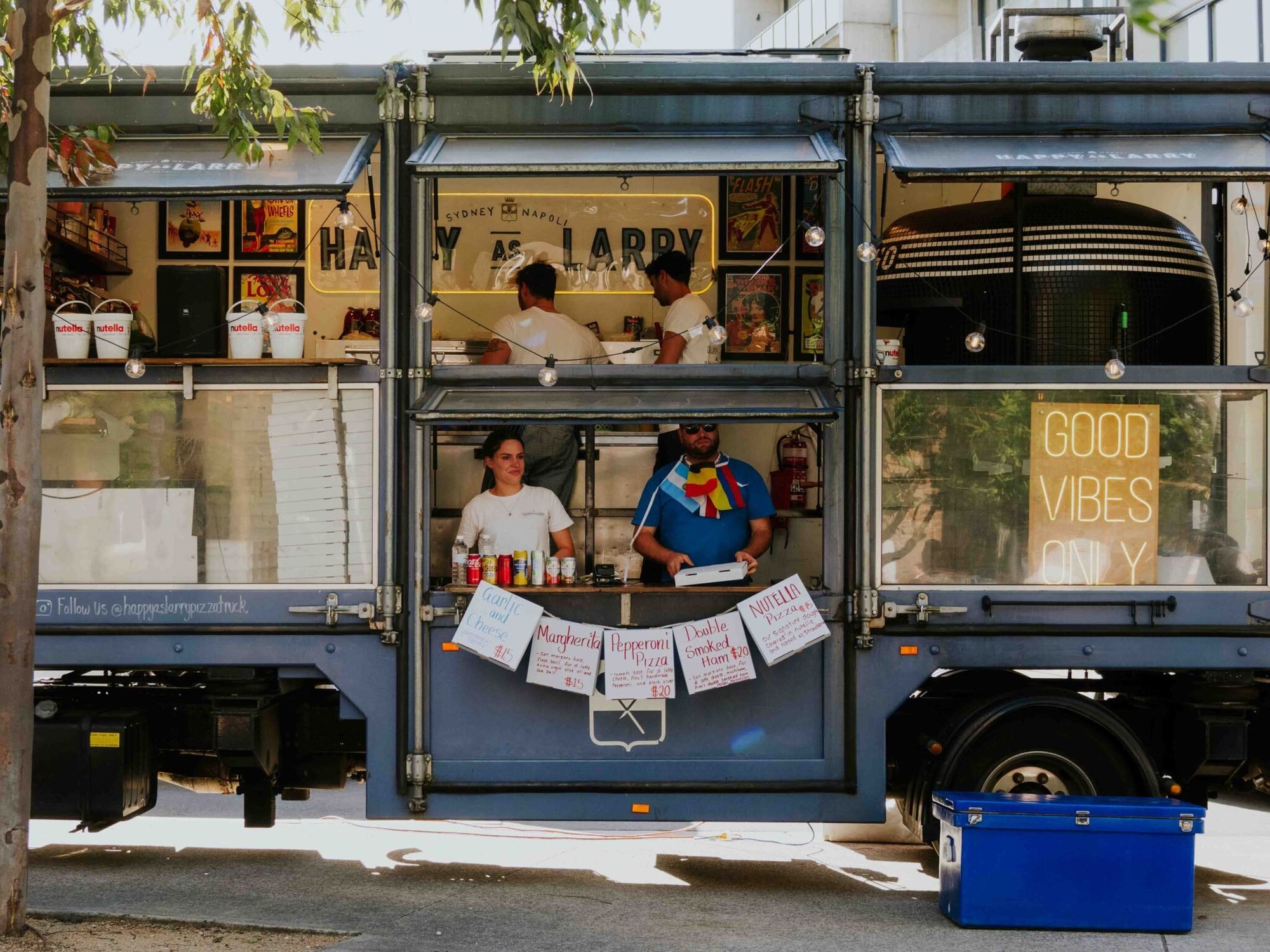 Food truck with "Good Vibes Only" sign, two vendors serving customers, colorful menu displayed, and a blue cooler on the ground.