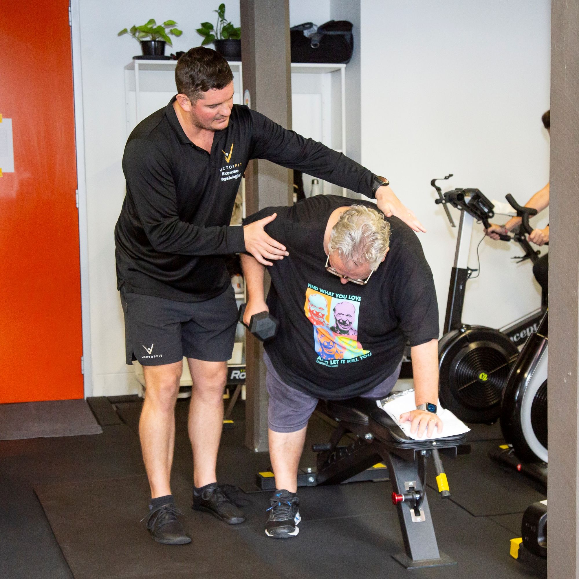 A trainer assists a person exercising with a dumbbell on a bench in a gym, with plants and exercise equipment in the background.