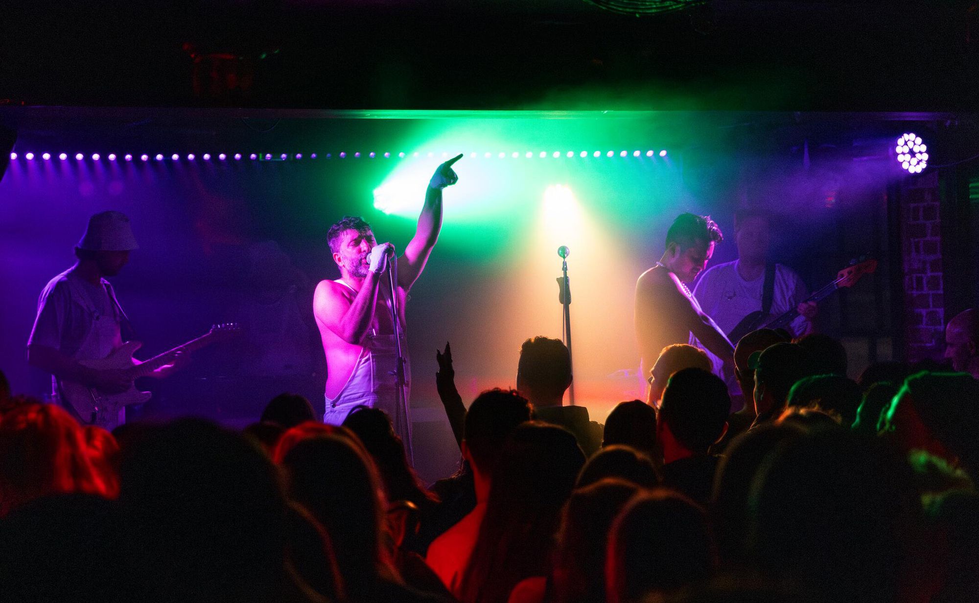 A band perform on stage in an intimate venue with a watching crowd in the foreground