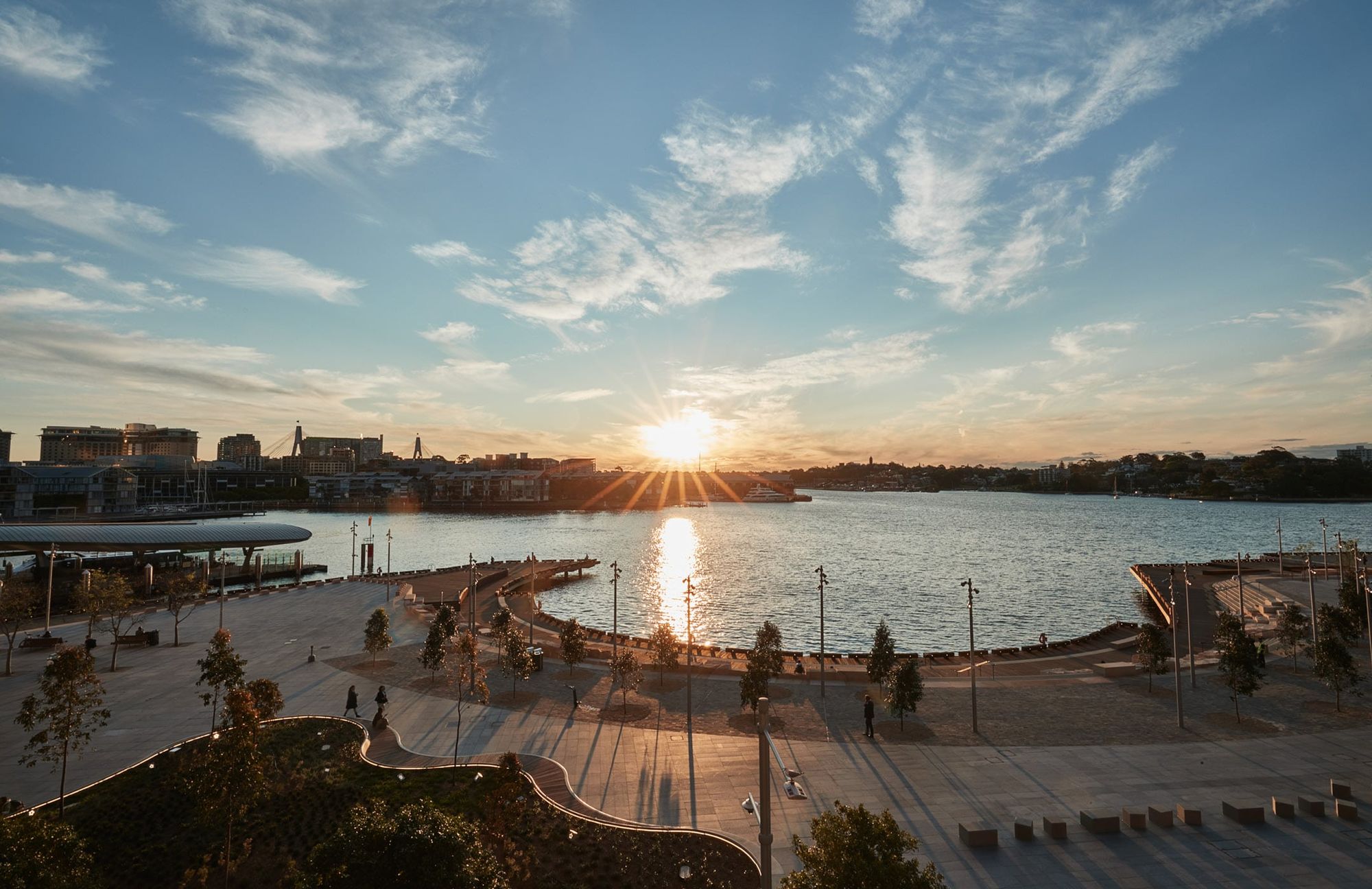 Sunset over a waterfront park with a walkway, scattered trees, and distant buildings. The sky is partly cloudy with sun rays reflecting on the water.
