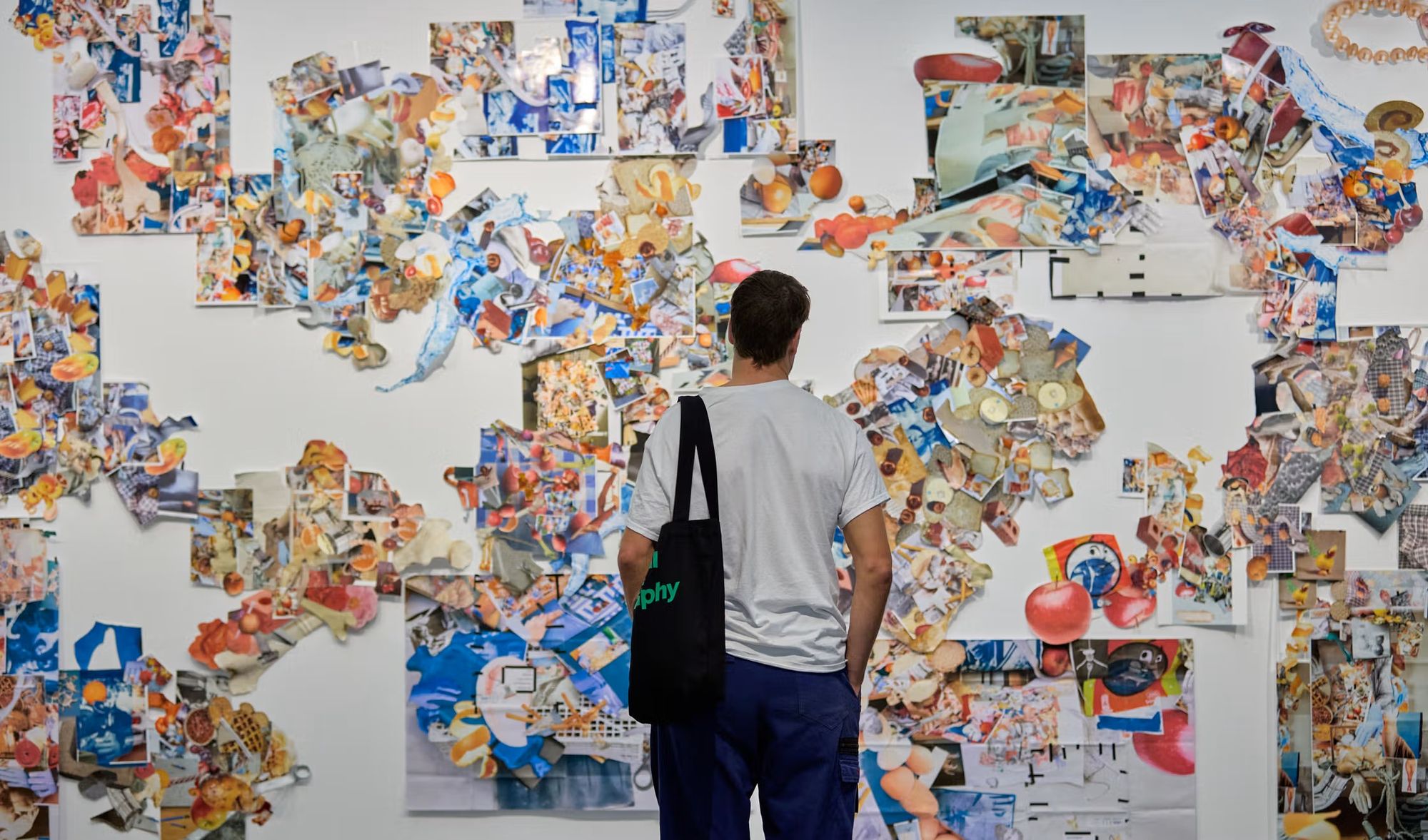 A person with a black bag observes a wall filled with colourful abstract art collages.