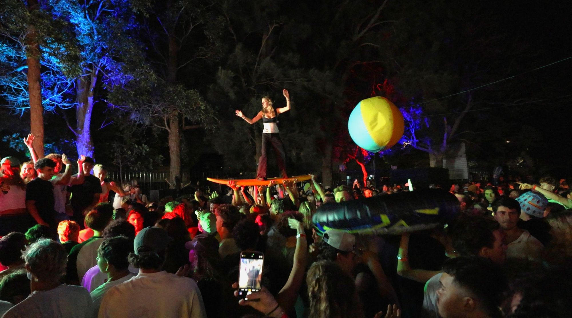 A woman crowd-surfing while standing on a surfboard held aloft by a cheering crowd  at a night-time outdoor festival, with a beach ball floating above the crowd.