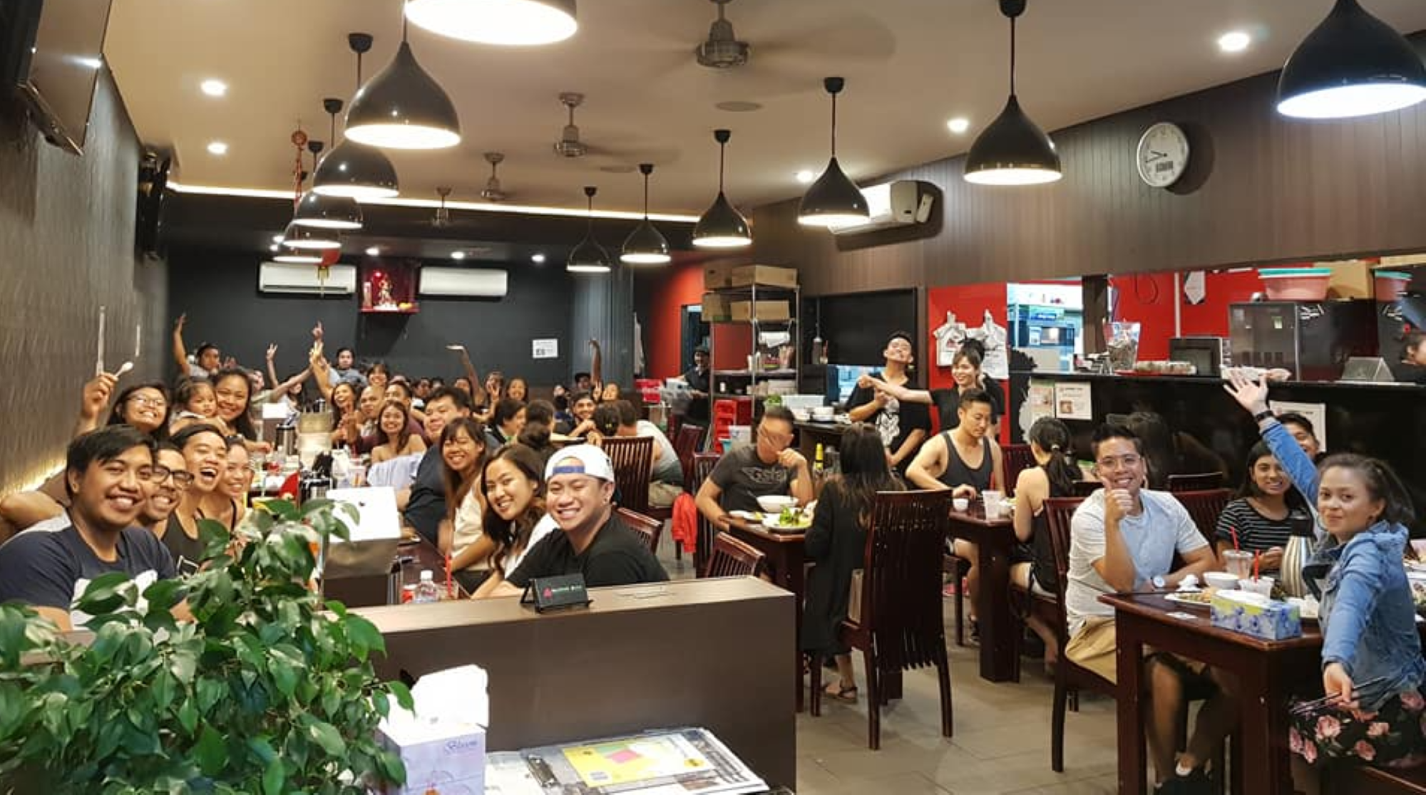 A large group of people smiling and posing for a photo in a lively restaurant with modern decor and hanging lights.