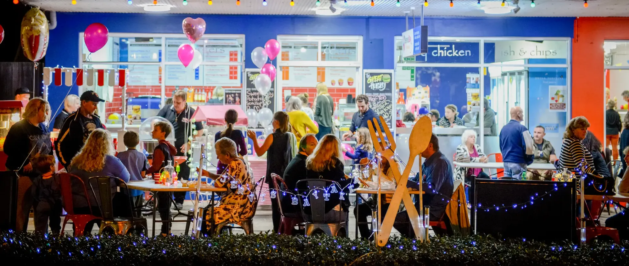 A lively nighttime scene of people dining outdoors at a brightly lit restaurant, decorated with balloons and string lights.