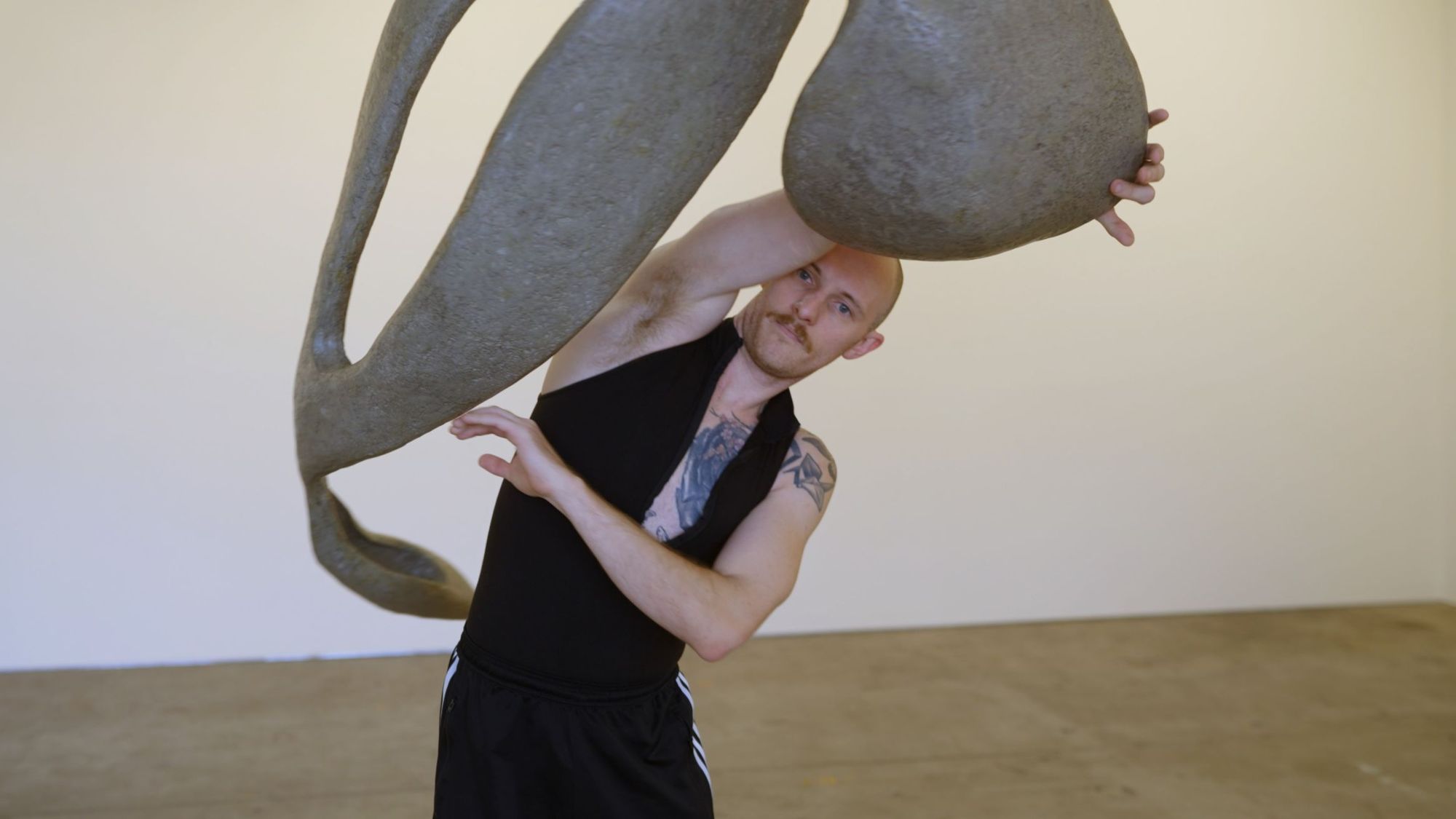 A person in a sleeveless top and shorts is posing with a large, abstract sculpture in a gallery space.