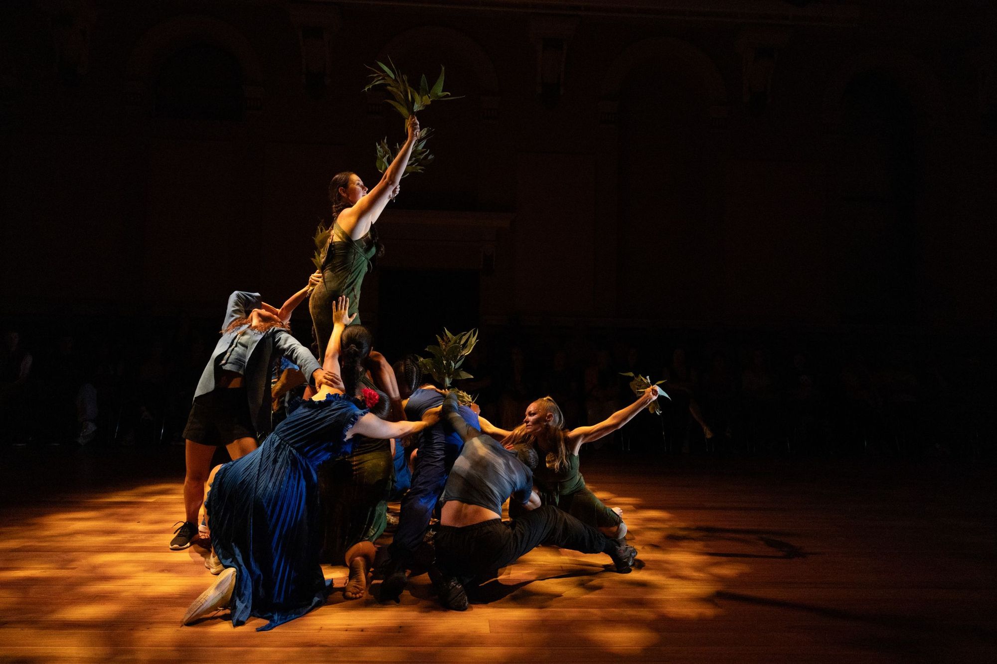 A group of performers form a dynamic, intertwined pose, with one standing atop, raising greenery under dramatic lighting on a wooden stage.