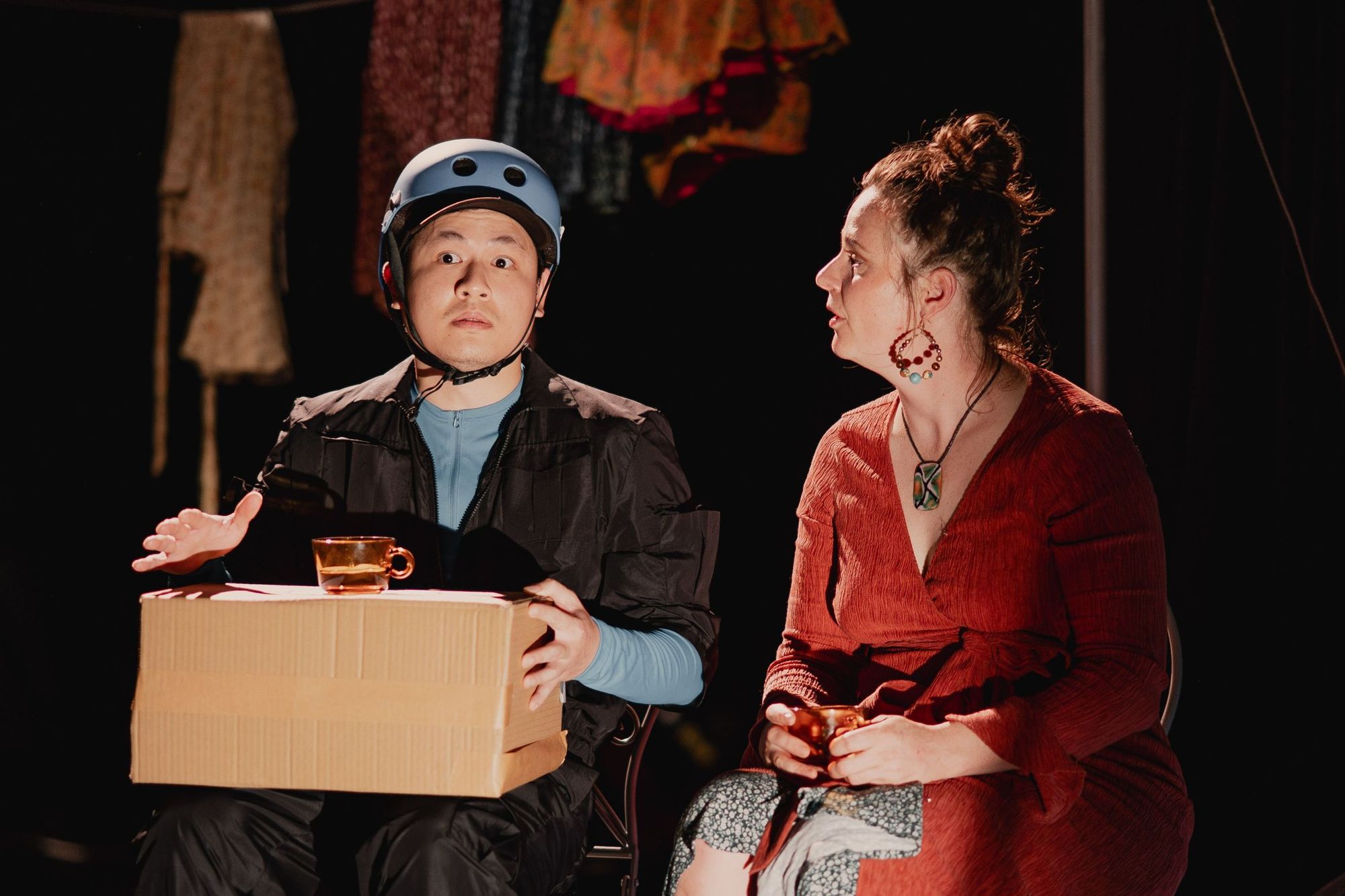 A person in a helmet holds a box and cup, looking surprised, while sitting beside another person in a red dress holding a cup and looking at them.