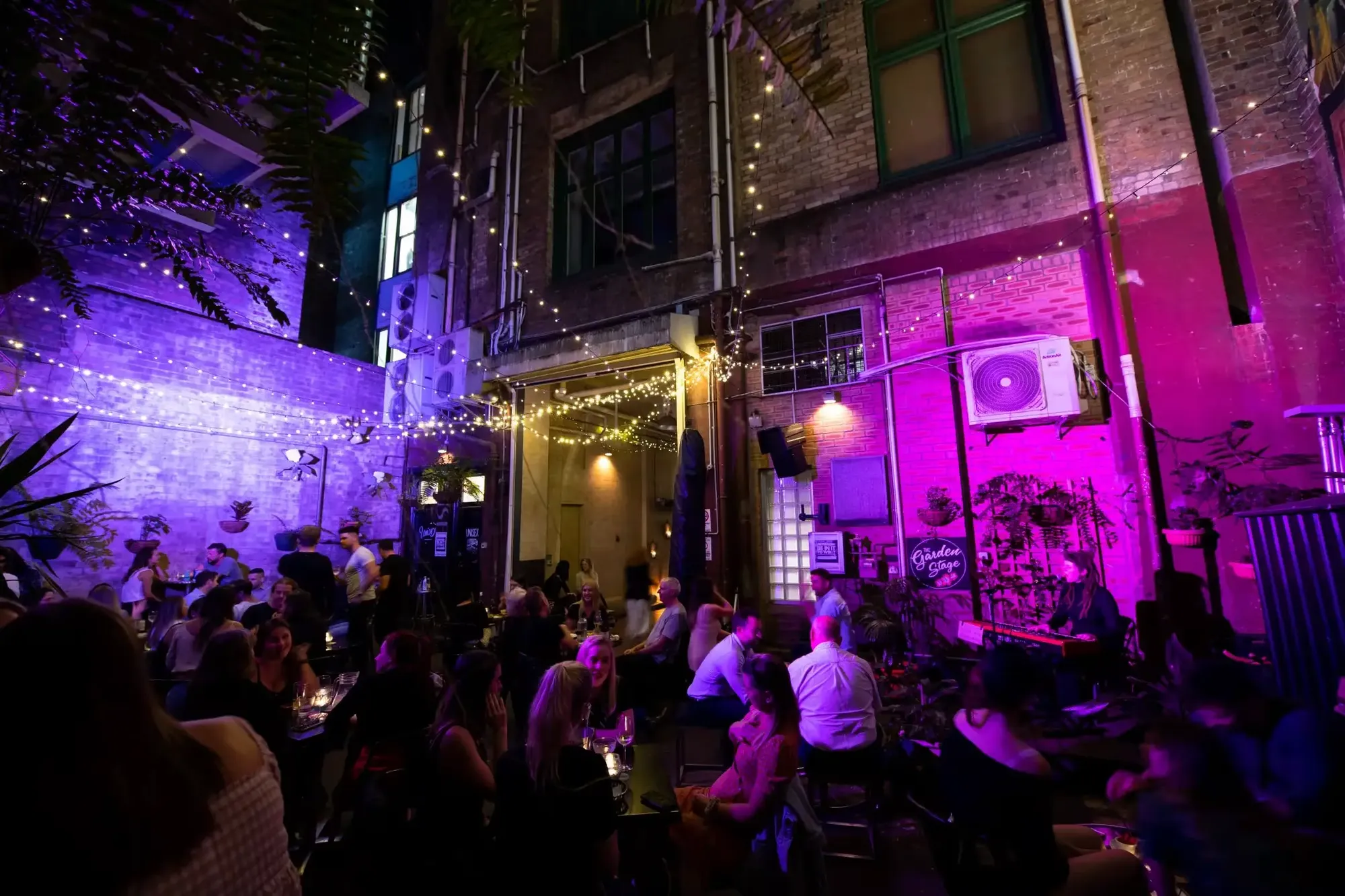 A vibrant courtyard bar with people seated at tables, surrounded by string lights and colorful purple and yellow lighting on brick walls.