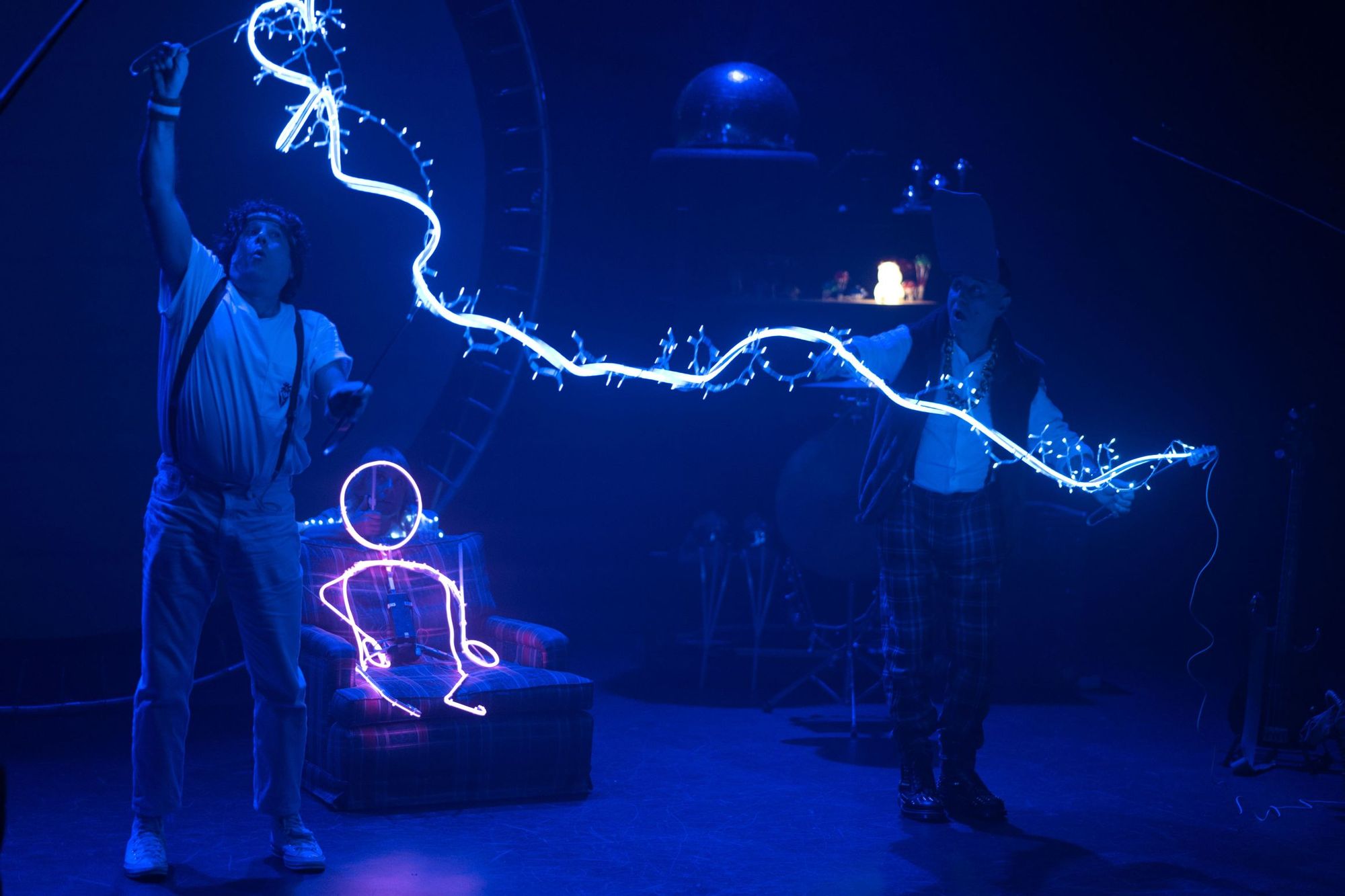 Two performers on stage with glowing blue lights; one holds a wand-like object creating glowing designs, while another manipulates wires.