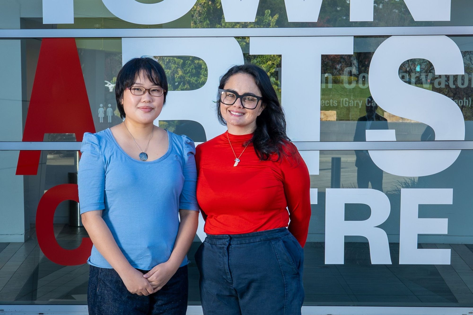 Two women stand smiling in front of a building with large glass windows displaying the words "ARTS CENTRE" in bold letters.