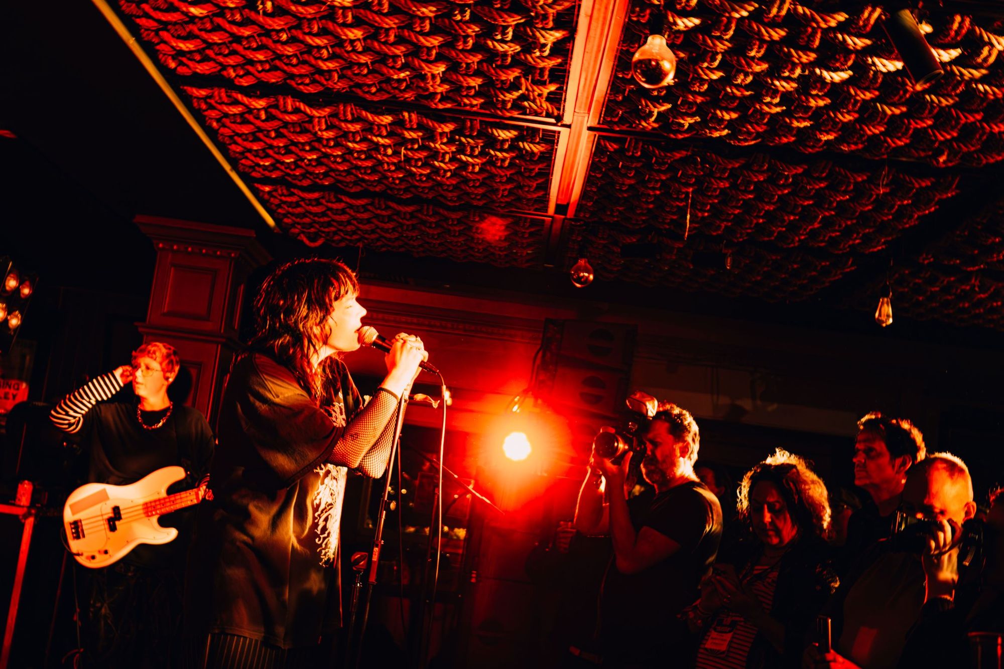 A singer performs passionately on stage, microphone in hand, with a guitarist beside them and an audience gathered under warm, dramatic lighting.