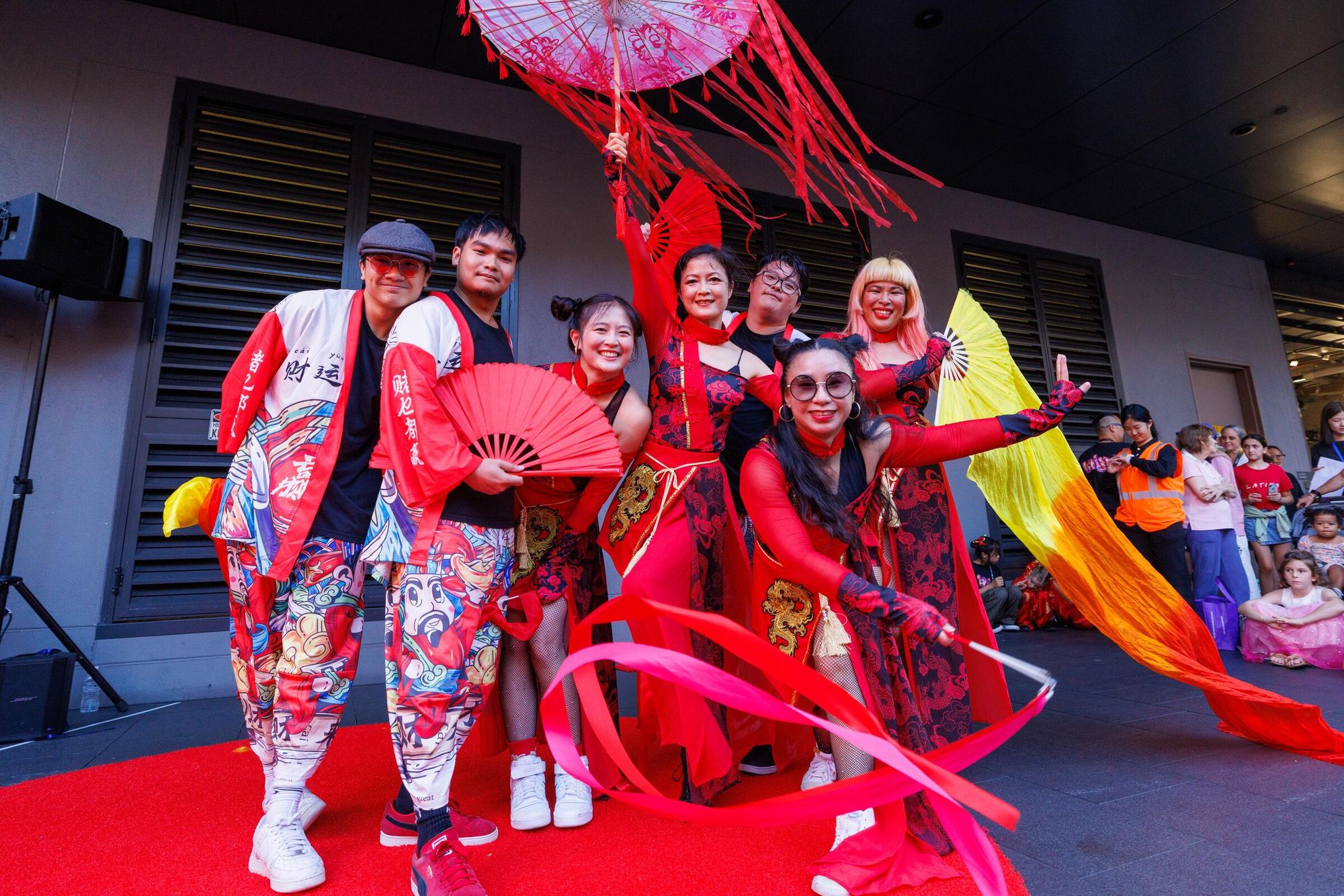 A group of dancers wearing contemporary Asian dance outfits smile to camera. They happily wave red ribbons, fans and streamers.