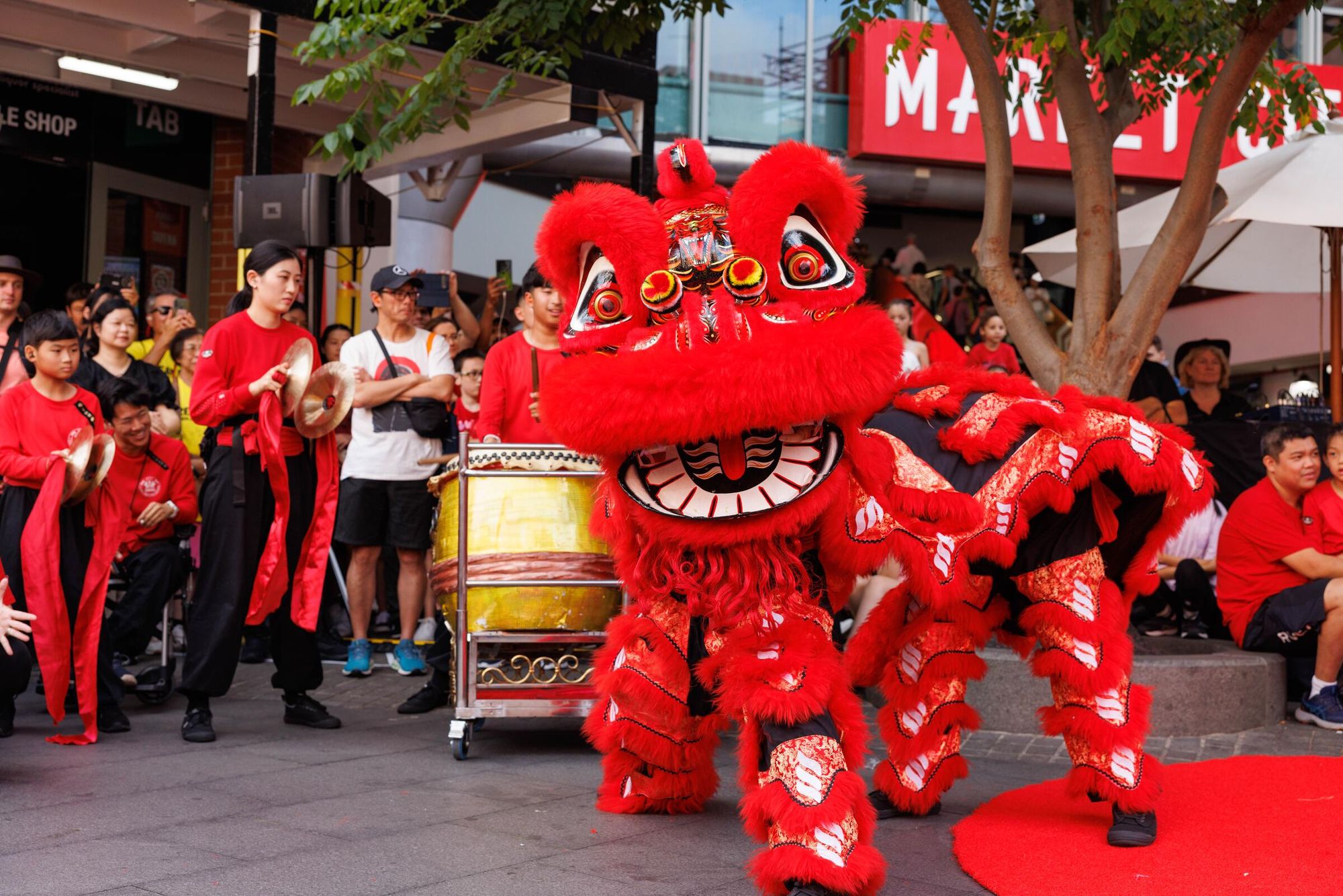 A red lion dancer performs on the street to a large crowd, supported by a drummer and two cymbal players.