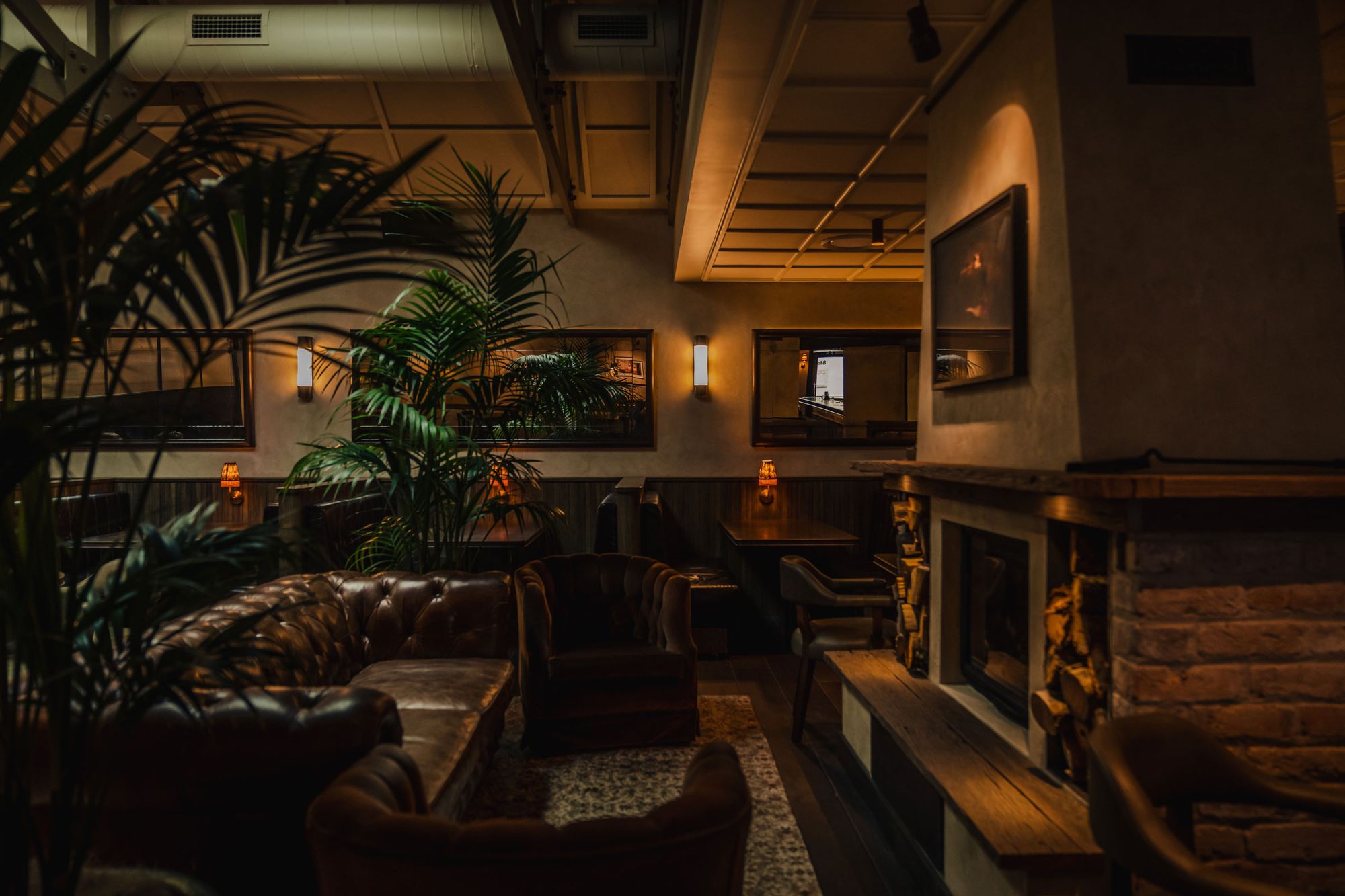 Cozy lounge with leather sofas, dim lighting, potted plants, and a brick fireplace, creating a warm and inviting atmosphere.