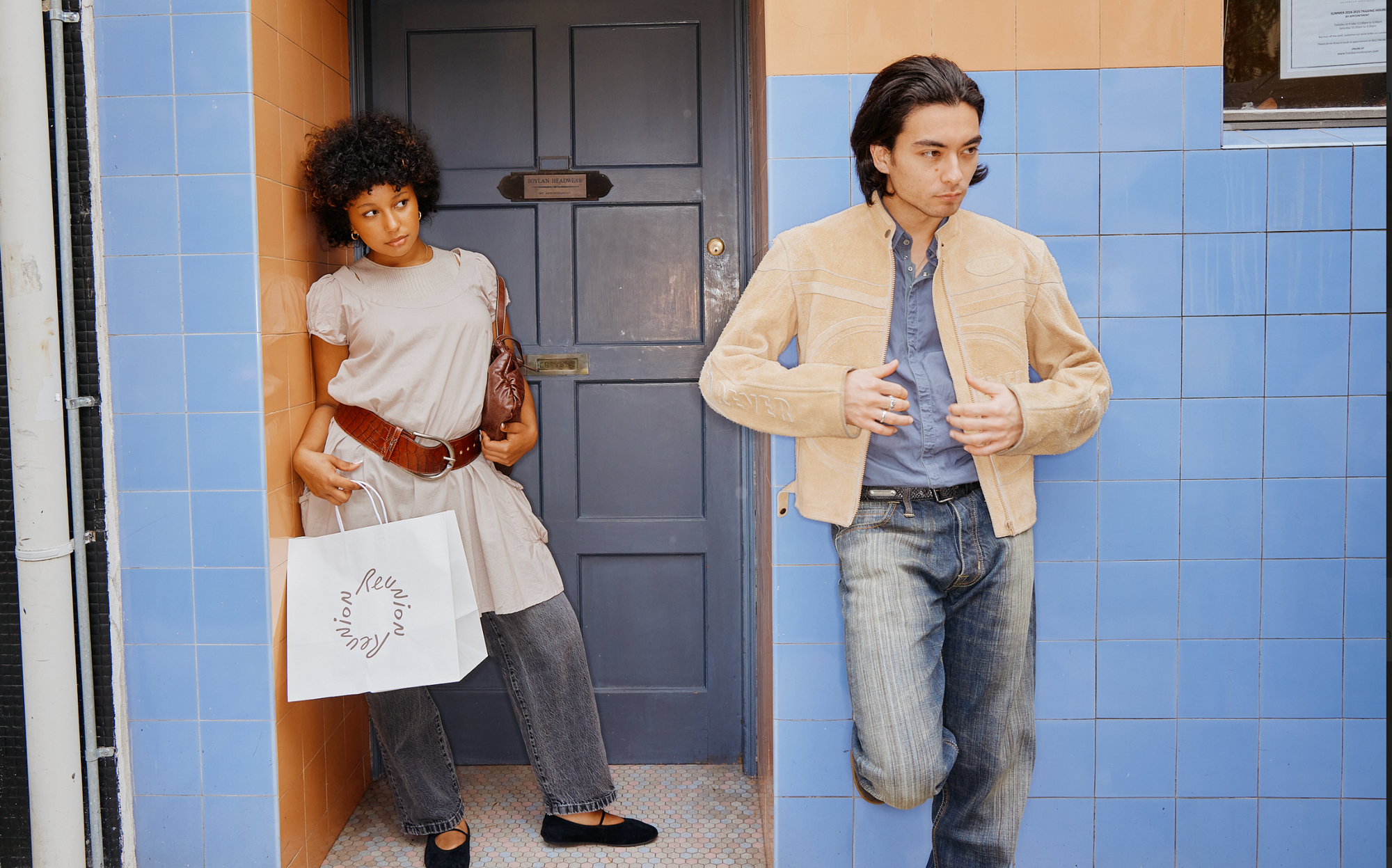 A woman stands in a doorway holding a shopping bag, while a man in a beige jacket walks past on a tiled street.