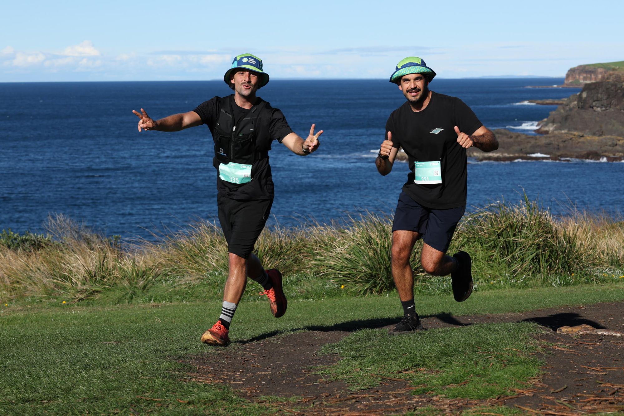 Two men in athletic gear and hats run joyfully along a scenic coastal path, with the ocean and cliffs in the background.