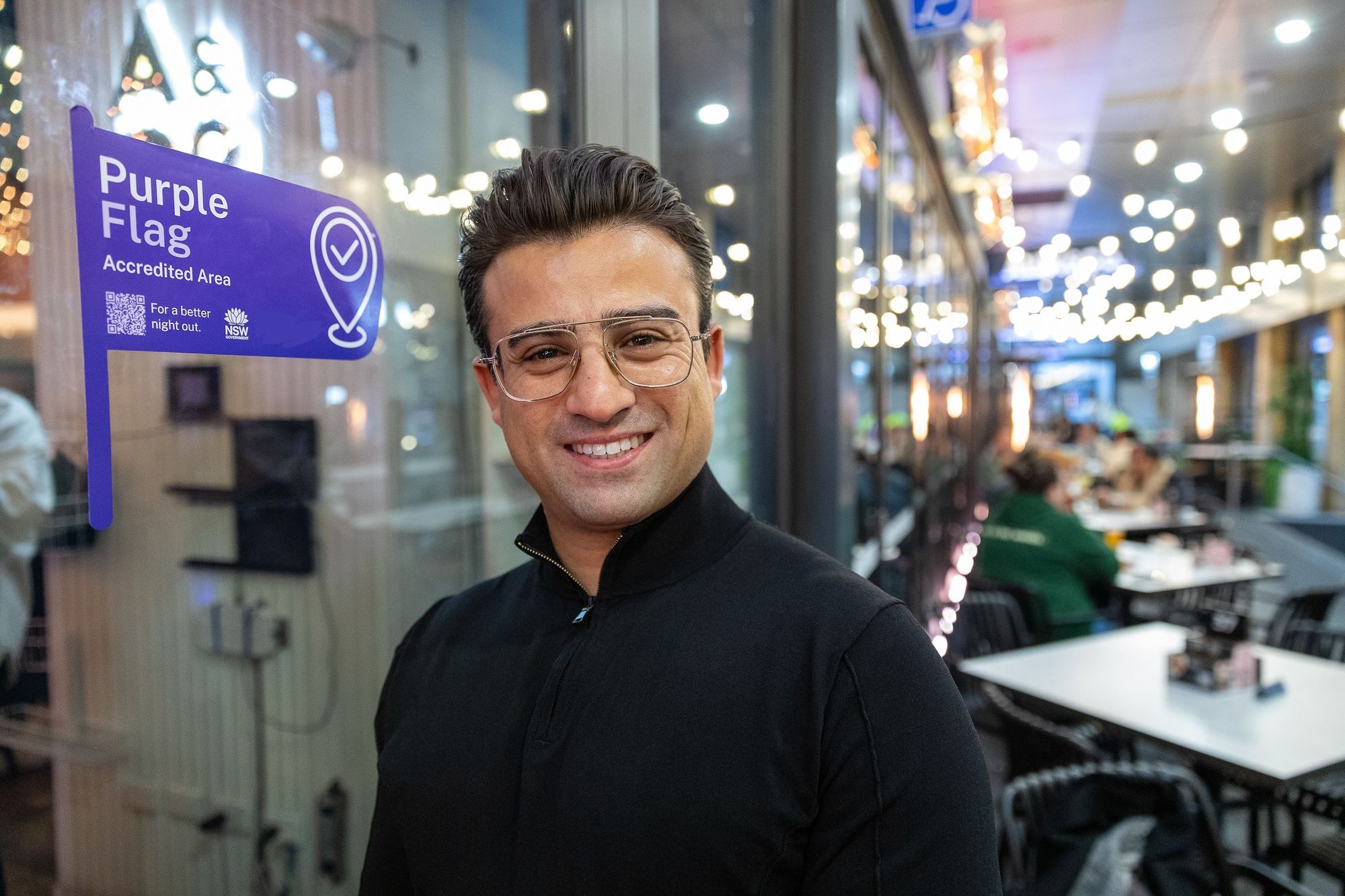 Smiling person in glasses stands by a "Purple Flag Accredited Area" sign, with a lively, well-lit outdoor dining area in the background.