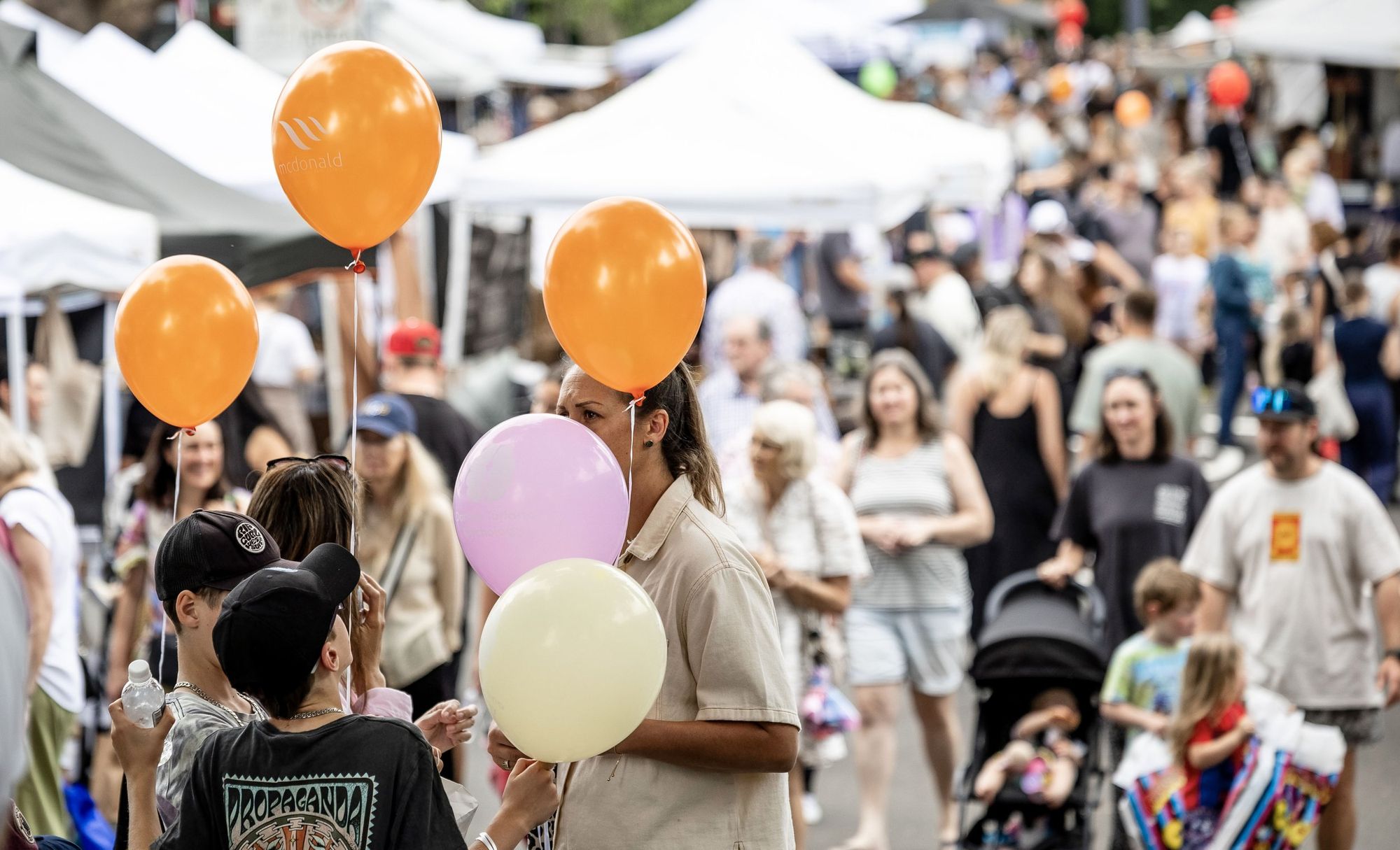 A crowded outdoor market with people holding colourful balloons, surrounded by tents and walking families on a bright day.
