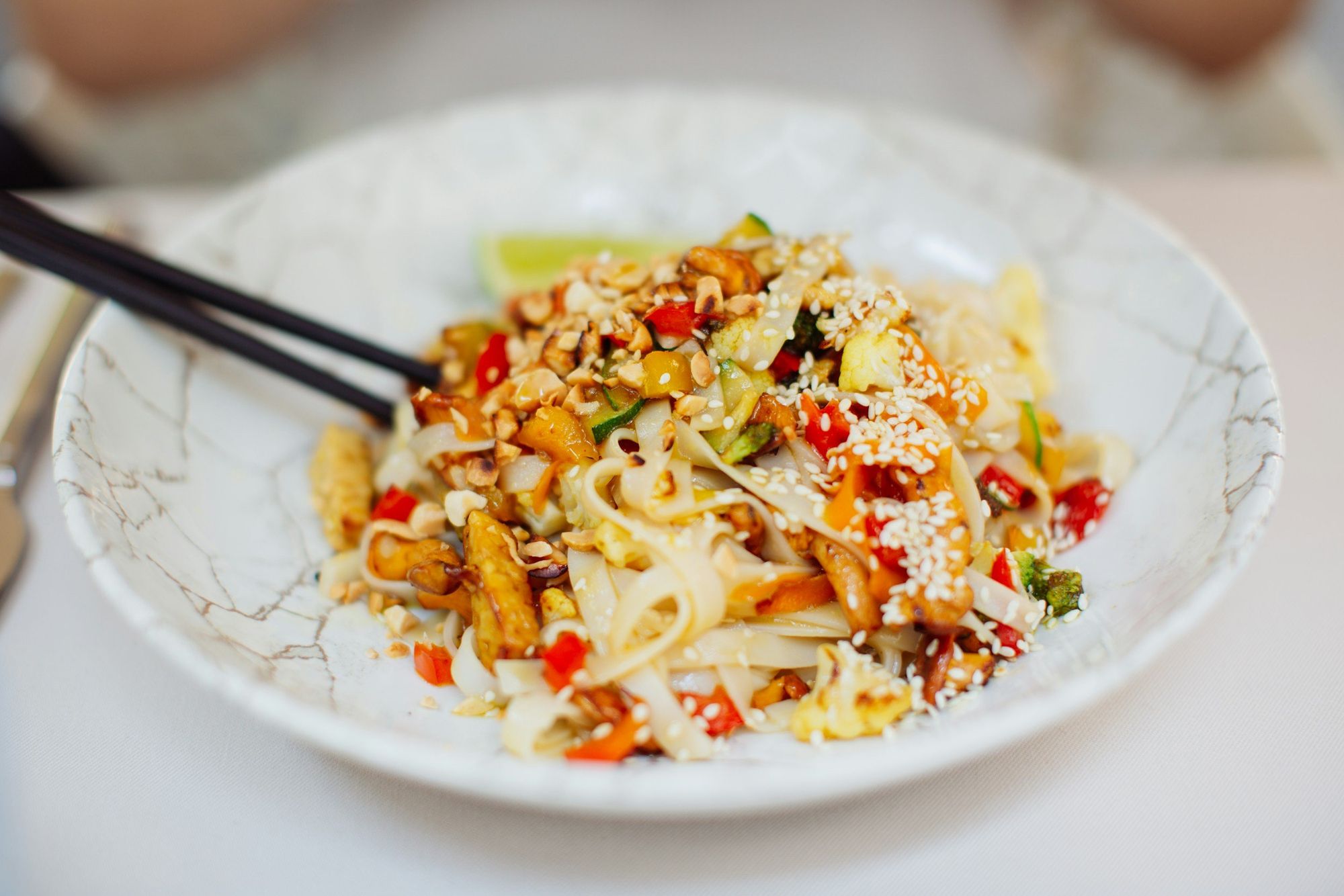 A dish of stir-fried noodles with vegetables, tofu, and sesame seeds, garnished with lime slice, served in a white bowl with chopsticks.