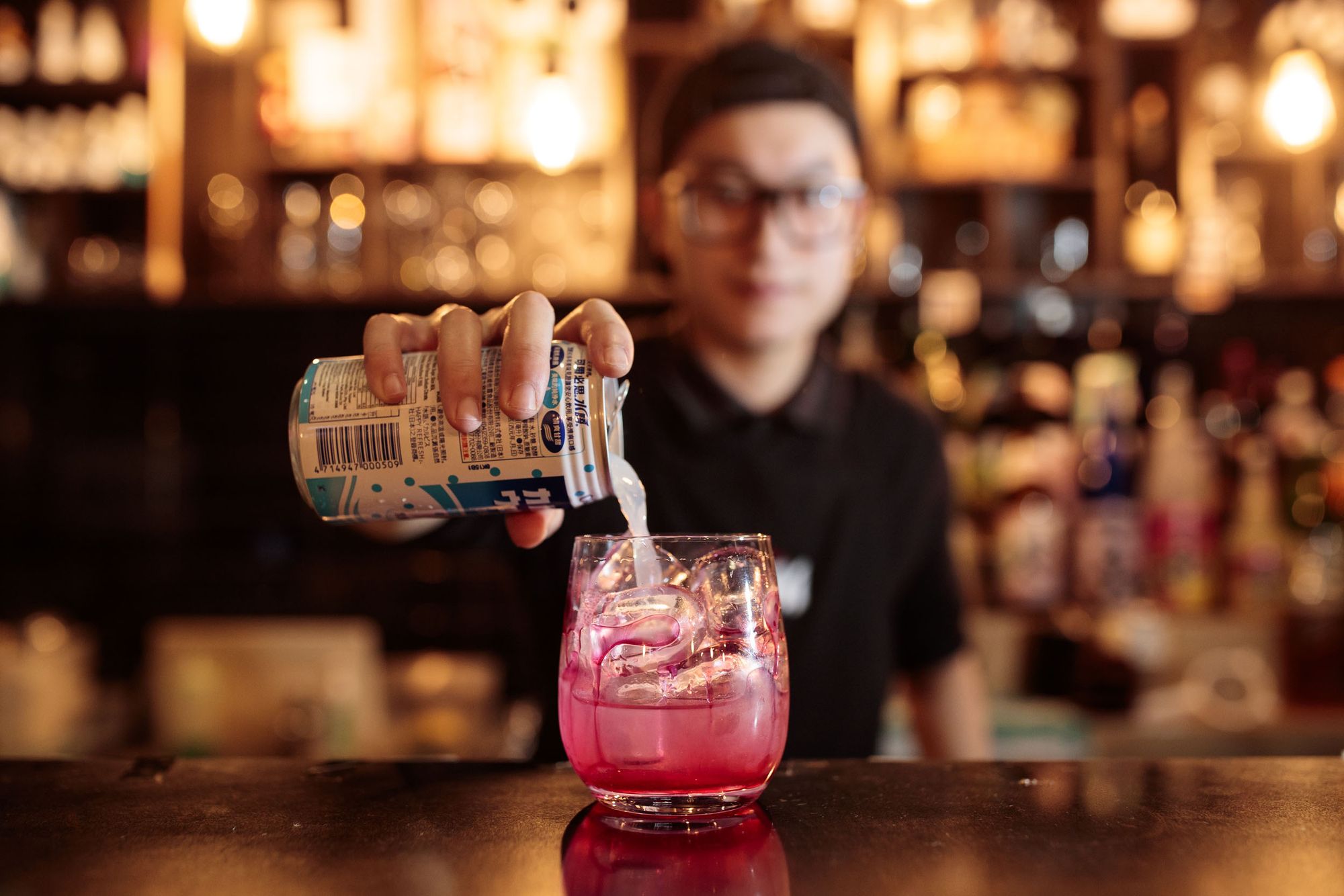 A bartender pours a drink from a can into a glass with ice, creating a pink beverage. The bar background is softly blurred.
