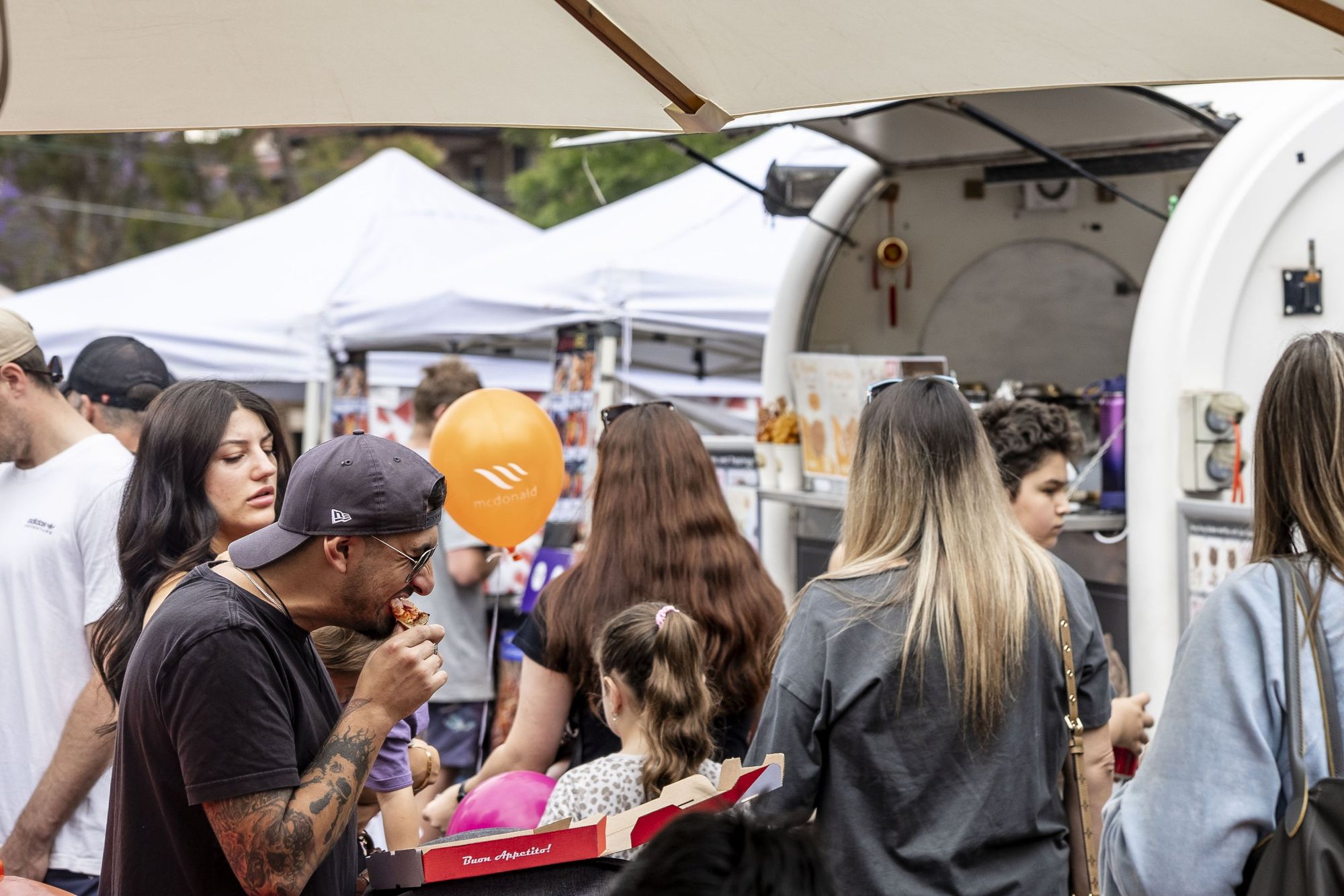 People at a bustling outdoor fair, some holding food and balloons, surrounded by white tents and a food truck.