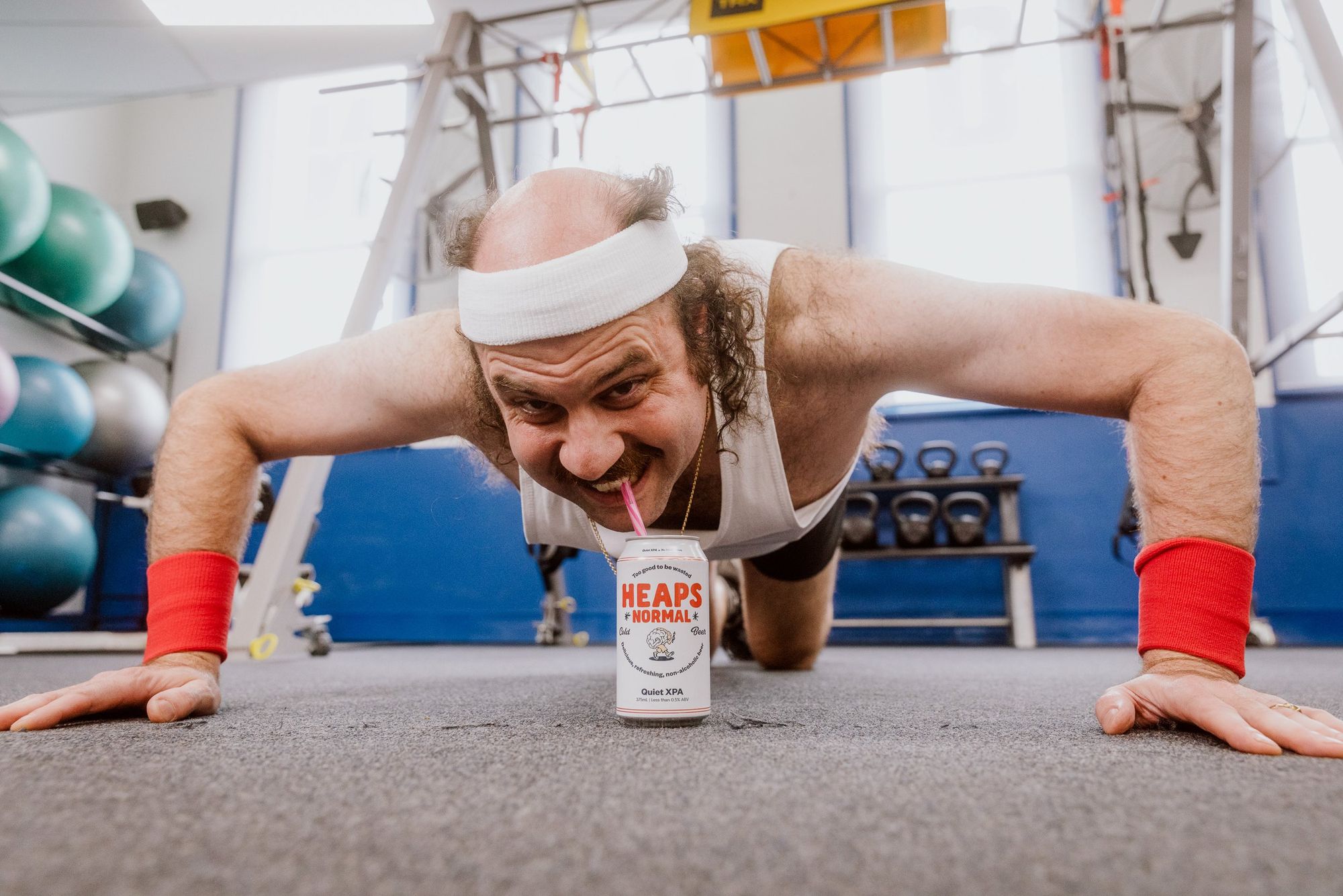 Man doing push-ups in a gym, wearing a headband and sipping from a can with a straw. Kettlebells and exercise balls in the background.