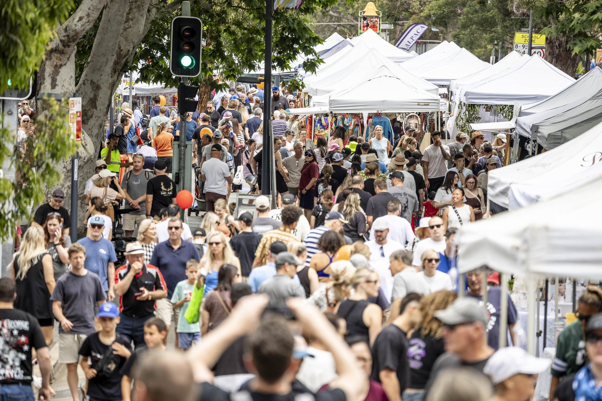 A bustling outdoor market with a large crowd, white tents, and a green traffic light in the background, surrounded by trees.