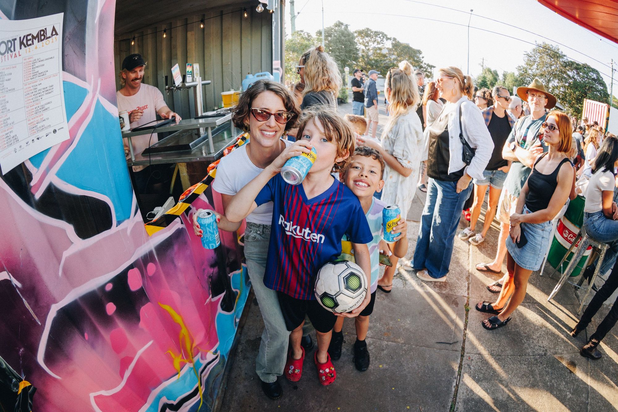 People enjoy drinks at an outdoor event, with a woman and two kids smiling by a colorful mural. One child holds a soccer ball.
