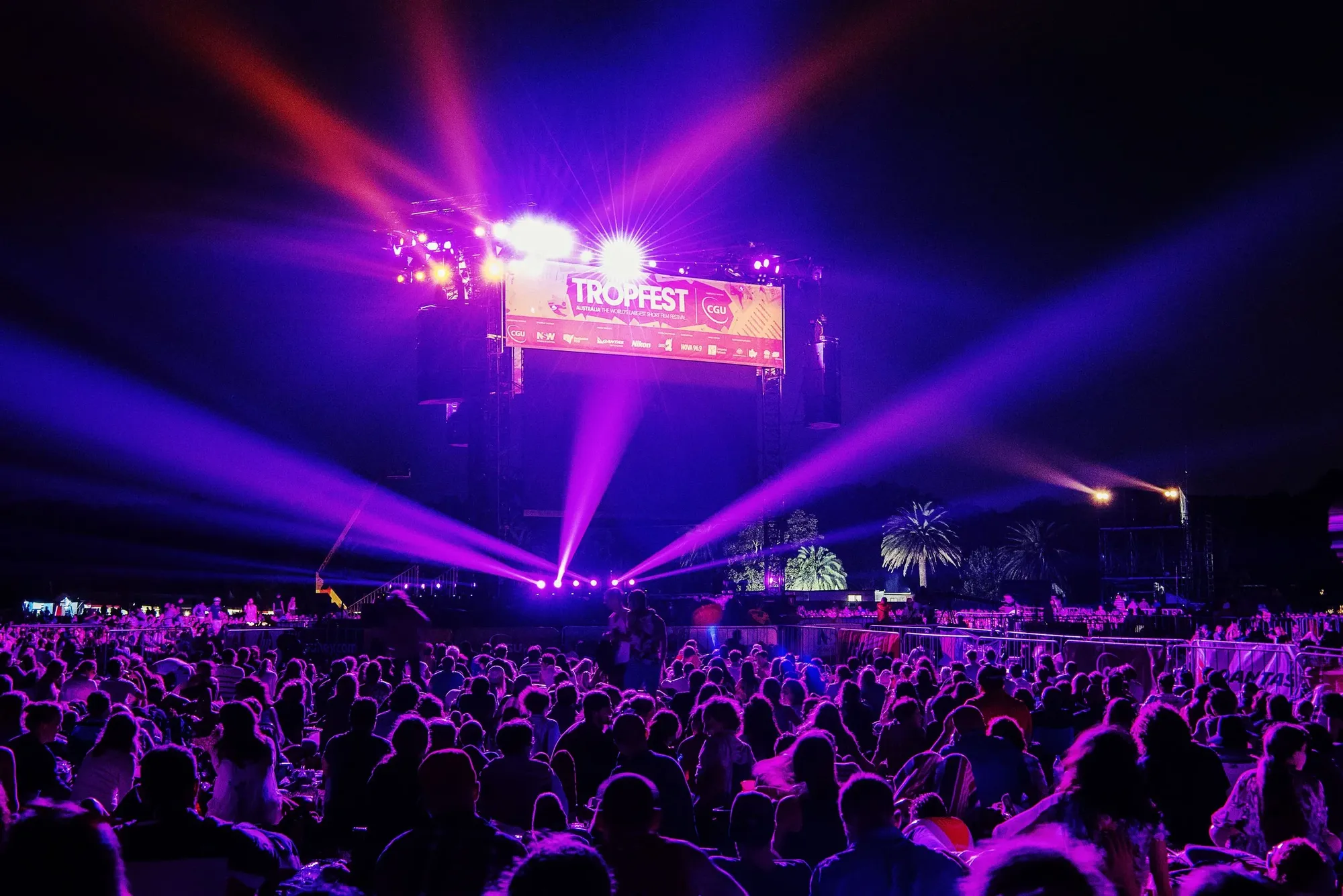 Large seated nighttime crowd watching a Tropfest stage with vivid purple and pink spotlights and laser beams.