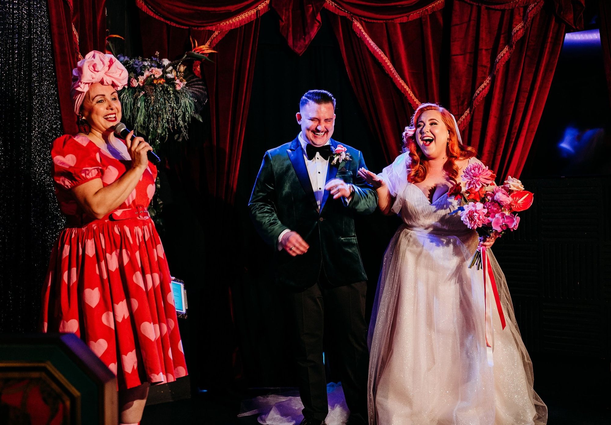 Three people on stage: a woman in a heart-patterned dress speaking, and a couple smiling, the bride holding a bouquet, with red curtains in the background.