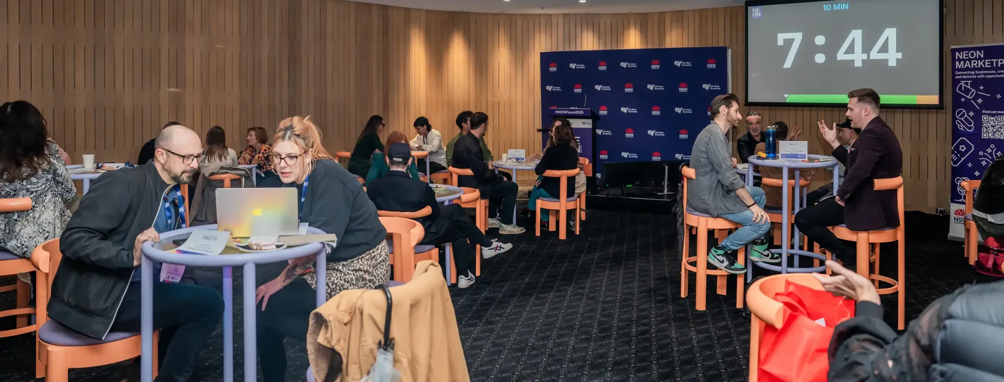 People sitting in groups at round tables, engaging in discussions; a timer on the wall shows 7:44.