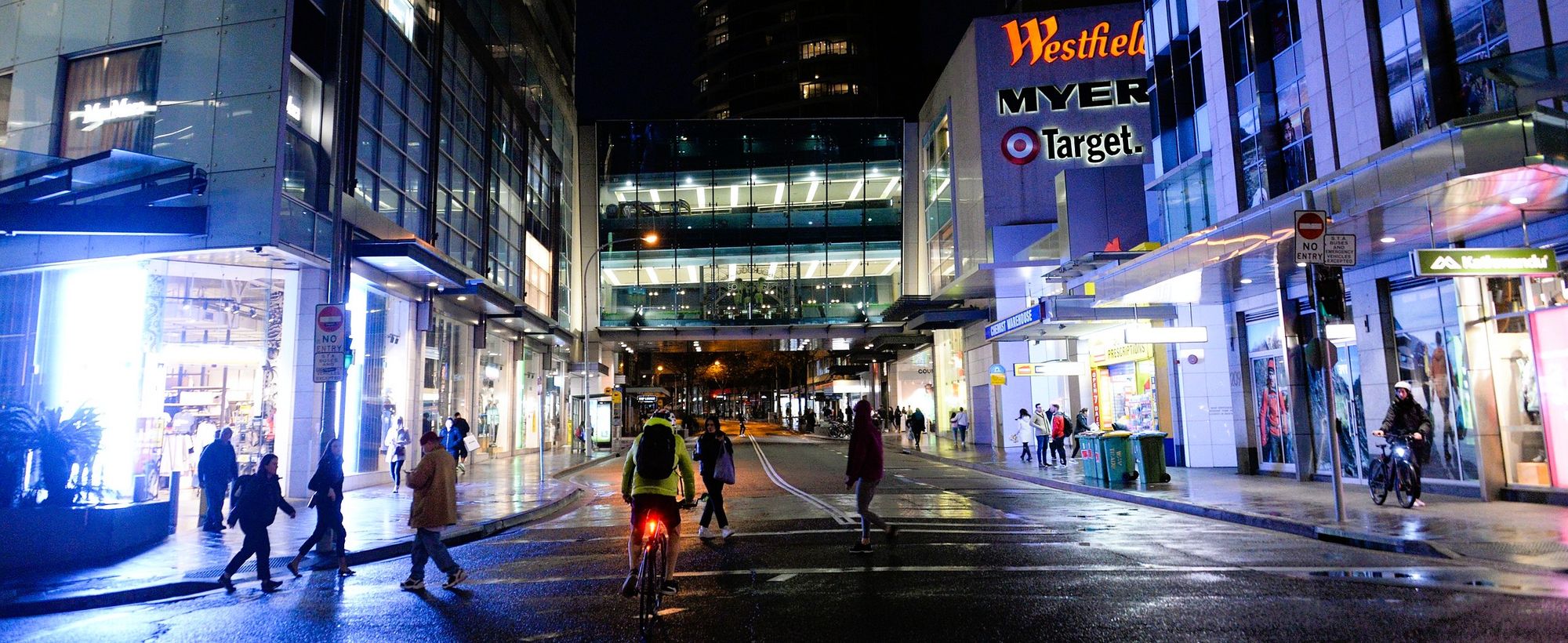 A busy urban street scene at night with pedestrians and a cyclist under bright store signs, including Westfield, Myer, and Target.