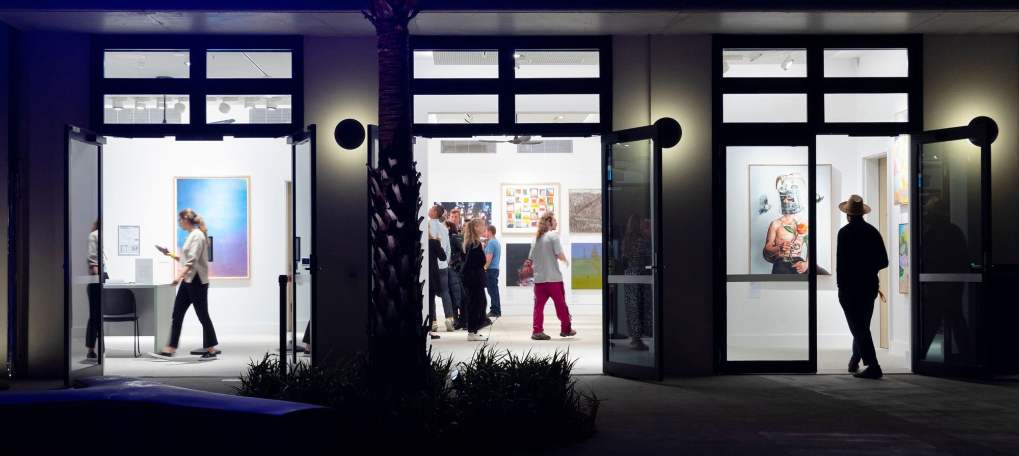 People browsing art in a brightly lit gallery with wide open doors, featuring various colorful paintings on the walls.