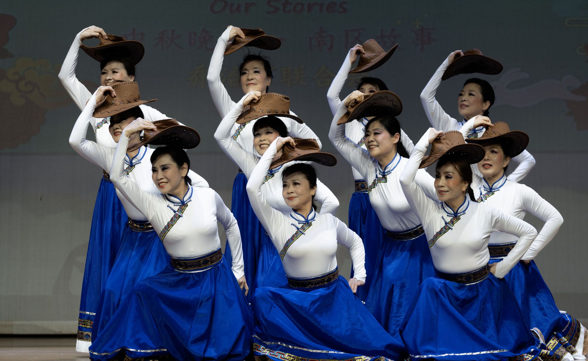 A group of performers in blue skirts and white tops pose with brown hats, smiling on stage during a cultural event.