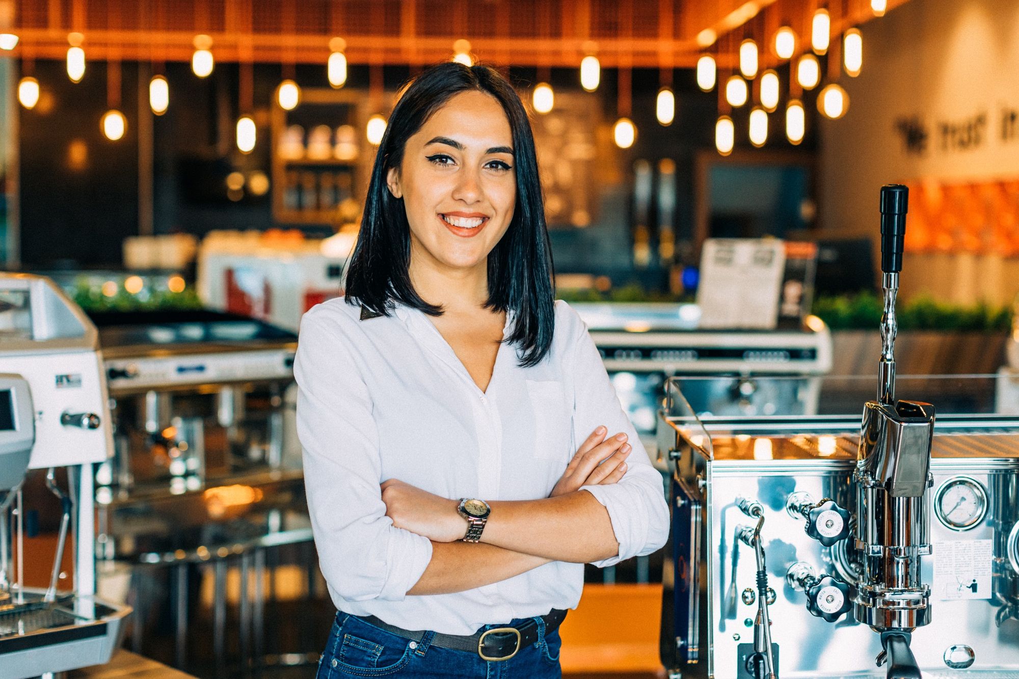 Smiling woman with crossed arms stands in a modern coffee shop, surrounded by espresso machines and contemporary lighting.