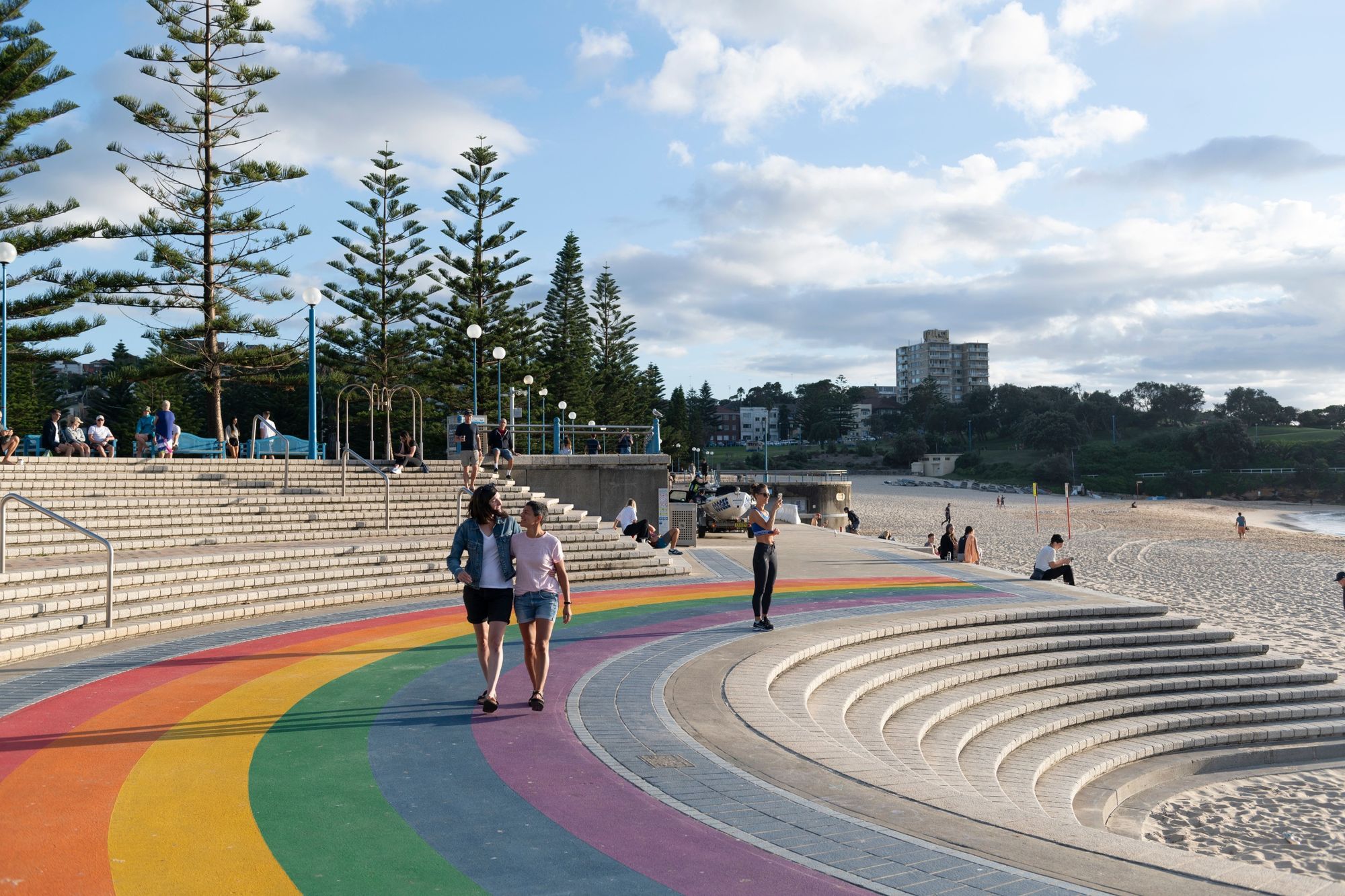 People walk along a colourful rainbow pathway by a beach, with steps and pine trees under a partly cloudy sky.