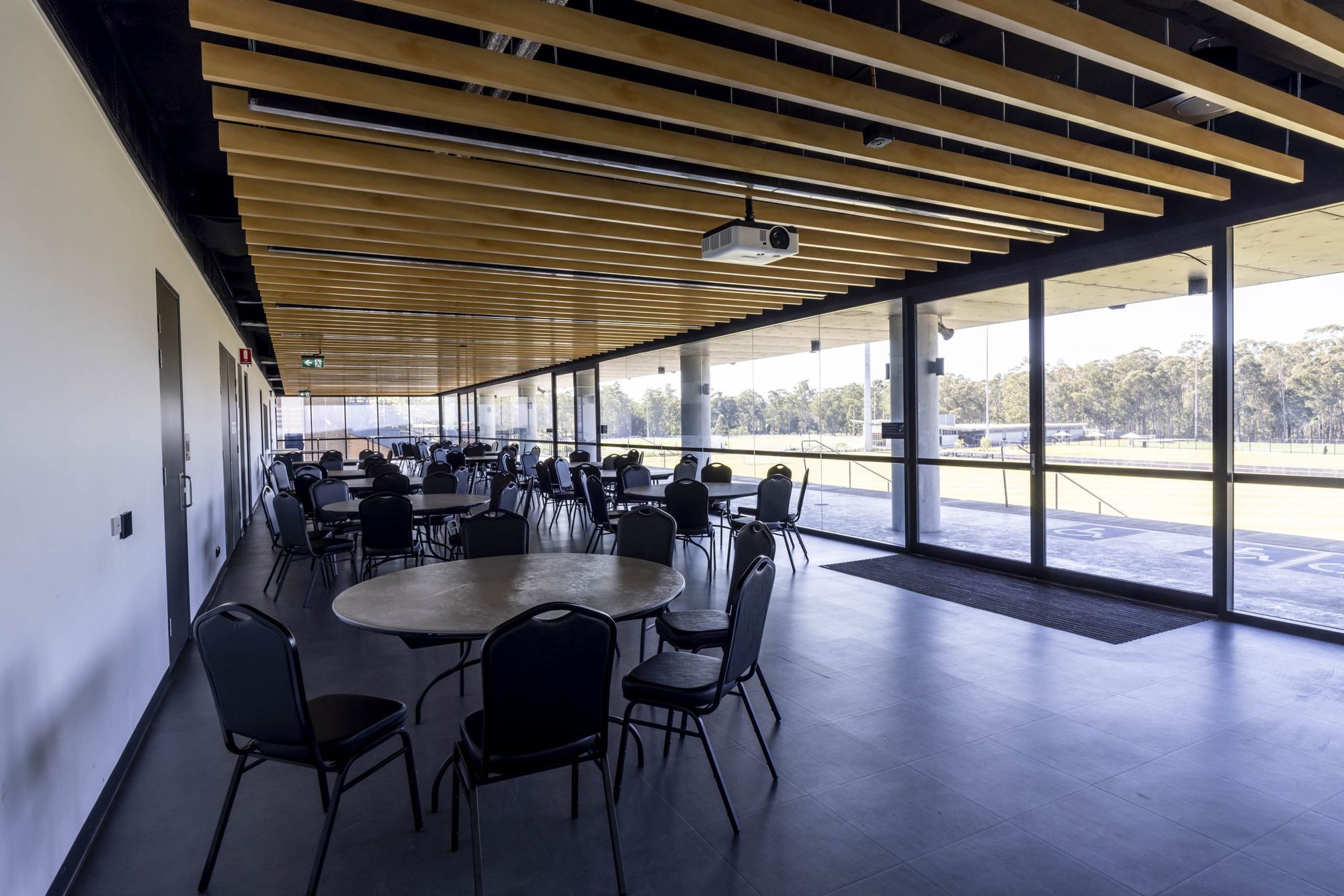 A spacious conference room with round tables and chairs, large windows, wooden ceiling panels, and a view of a sports field outside.