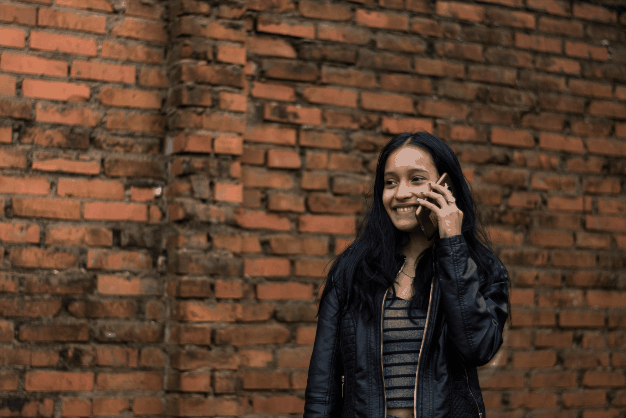 Smiling woman in a black leather jacket talks on the phone while standing in front of a brick wall.
