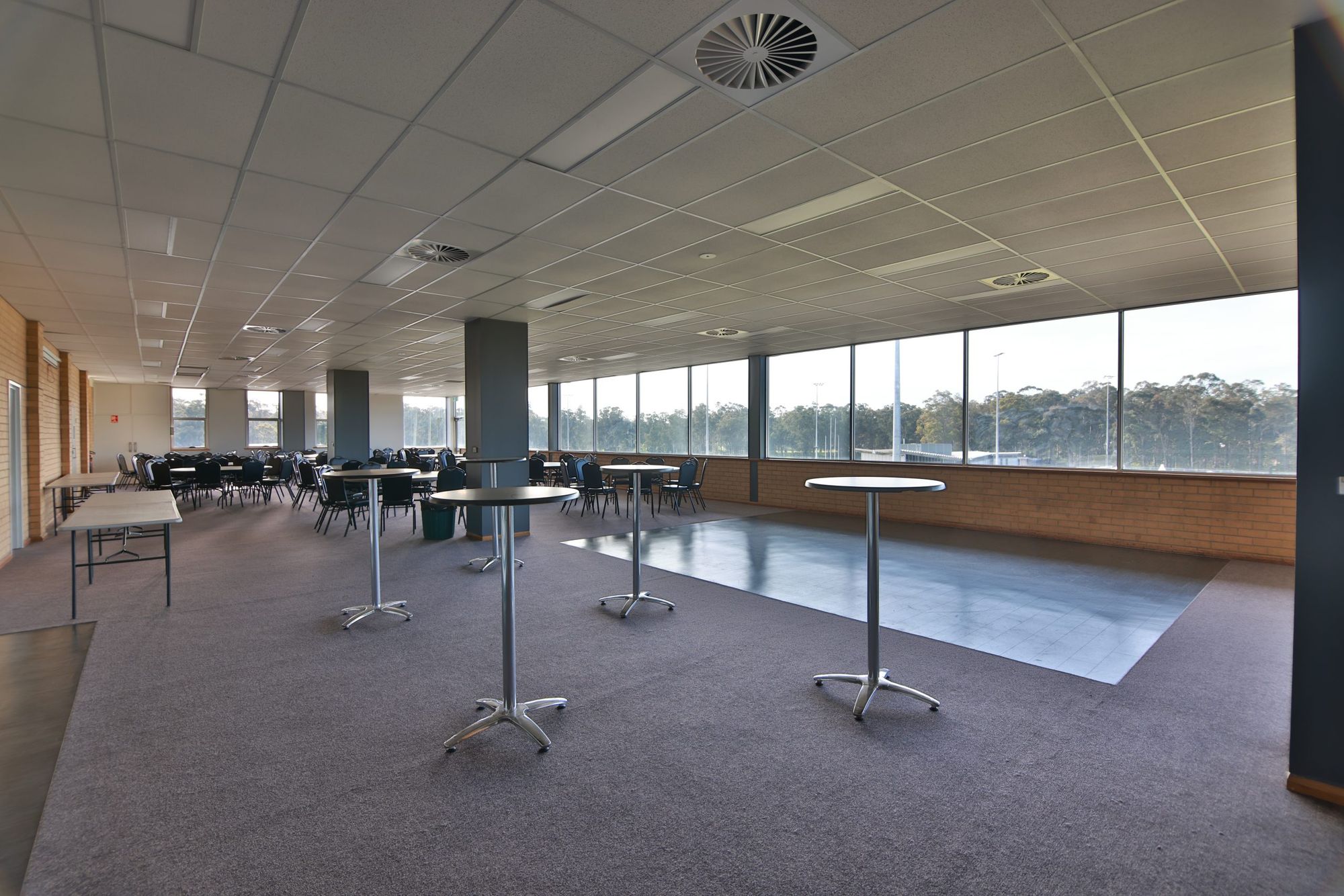 Spacious conference room with large windows, round tables, and chairs. Bright natural light illuminates the carpeted floor and minimalist decor.