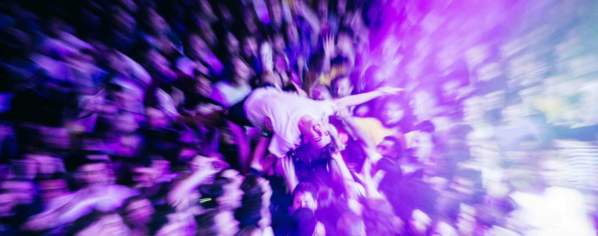 Person crowd surfing at a concert, surrounded by a vibrant, cheering audience under purple lighting.