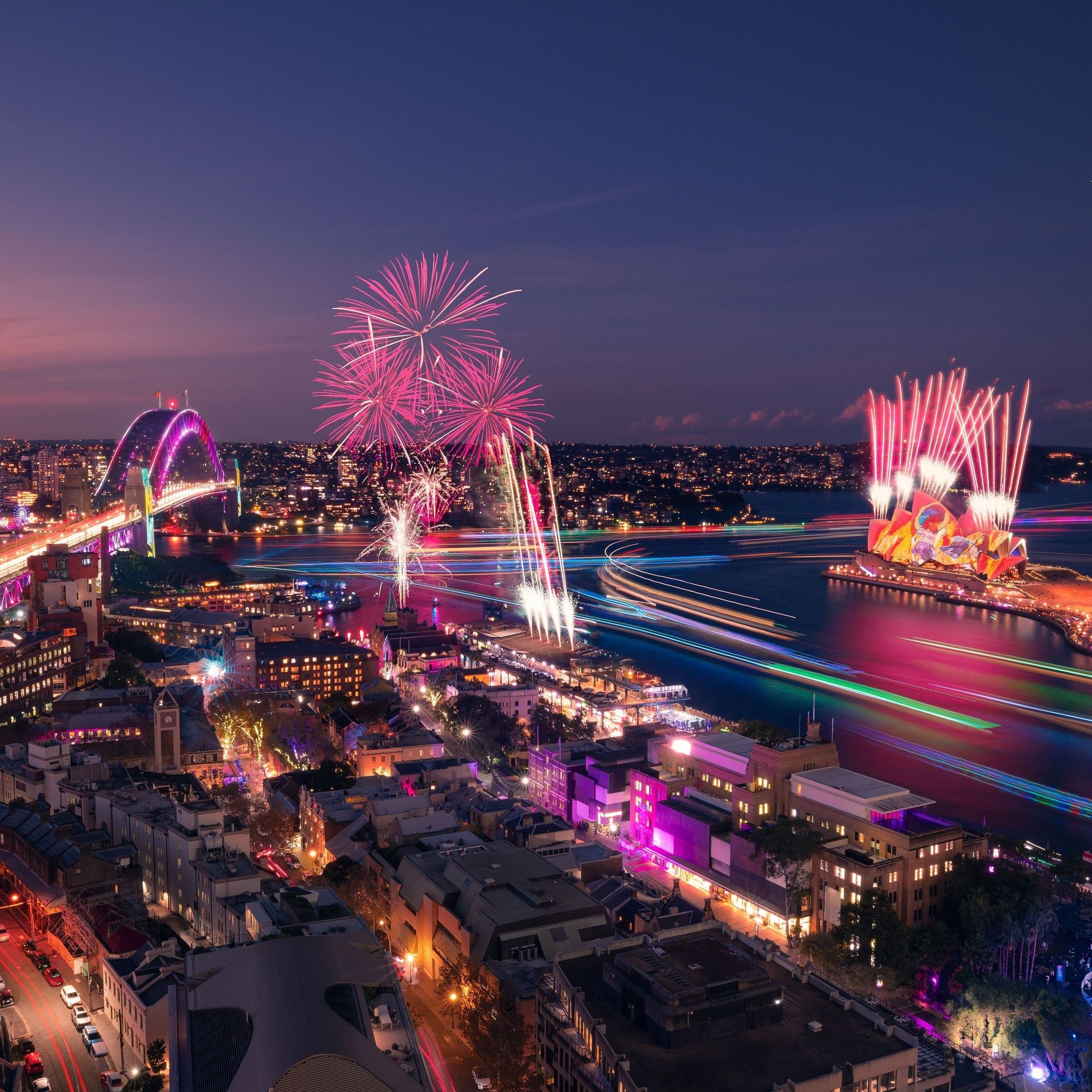 This nighttime image captures the magic of Vivid Sydney, with fireworks bursting in pink and white above the harbor. The Sydney Opera House comes alive with vibrant projections, while the Harbour Bridge glows in dynamic lights. The cityscape is illuminated with neon and LED displays, and light trails from boats streak across the water in shades of blue, green, and purple. The combination of art, technology, and celebration transforms Sydney into a breathtaking spectacle of color and movement.