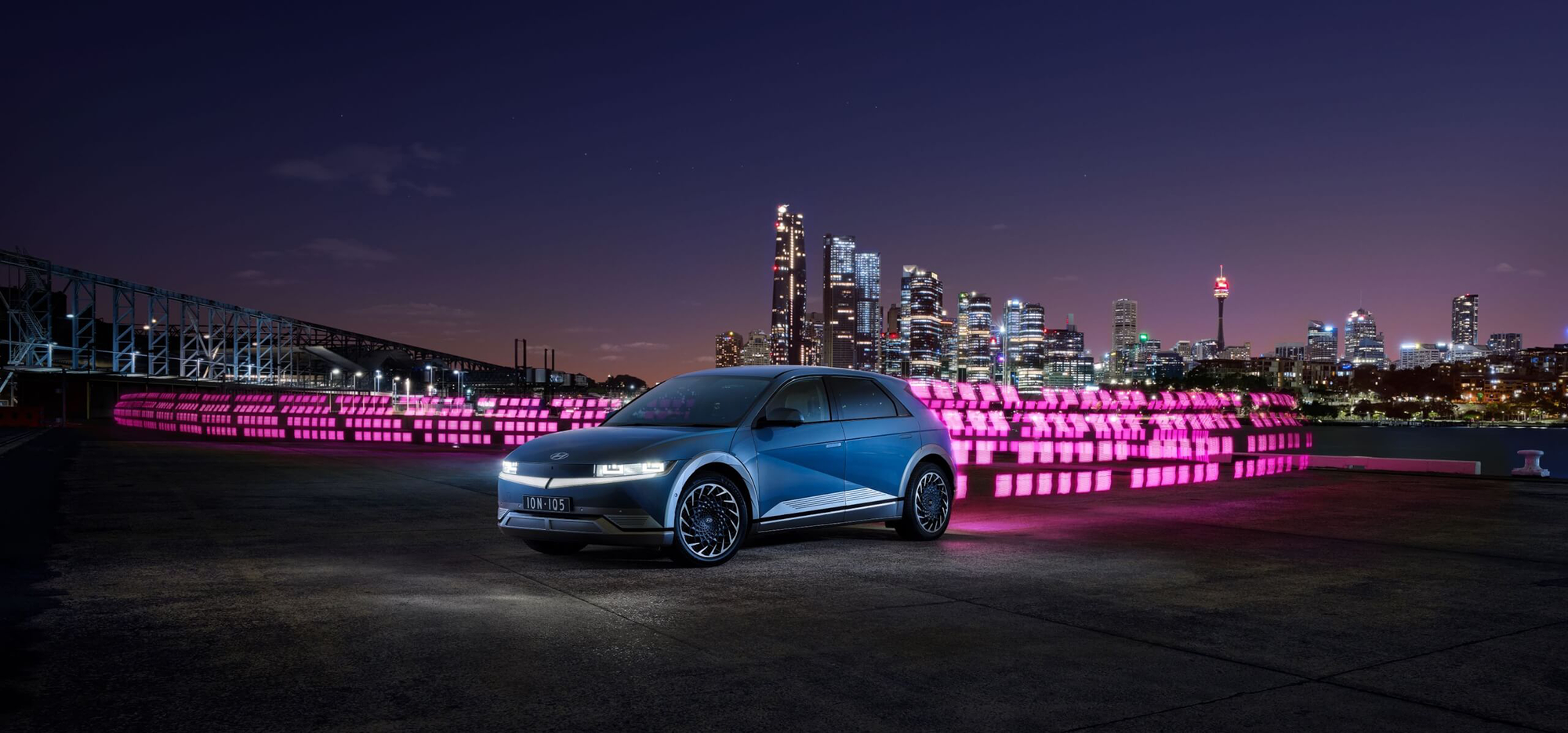 A wide cinematic night-time TVC production still featuring the Hyundai IONIQ 5 electric vehicle parked on a dark harbour-side concrete surface with the full Sydney CBD skyline glowing at dusk behind it. The car is shot in a cool steel-blue finish with its headlights on, NSW number plate reading "ION·IQ5". Stretching horizontally behind and around the vehicle is an elaborate custom light painting installation — hundreds of individually pixel-controlled magenta and hot-pink LED panels arranged in dynamic grid and wave patterns that sweep across the entire mid-ground, wrapping the car in vivid colour and creating a bold, designed landscape of light between the vehicle and the skyline. The Sydney Tower, Harbour Bridge approach structure, and illuminated city towers are clearly visible against the deep purple-blue twilight sky. The light painting technique was executed by Lightology Australia in collaboration with Eat the Elephant and Scoundrel Films, purpose-built and captured in-camera for the Hyundai IONIQ Range TVC.