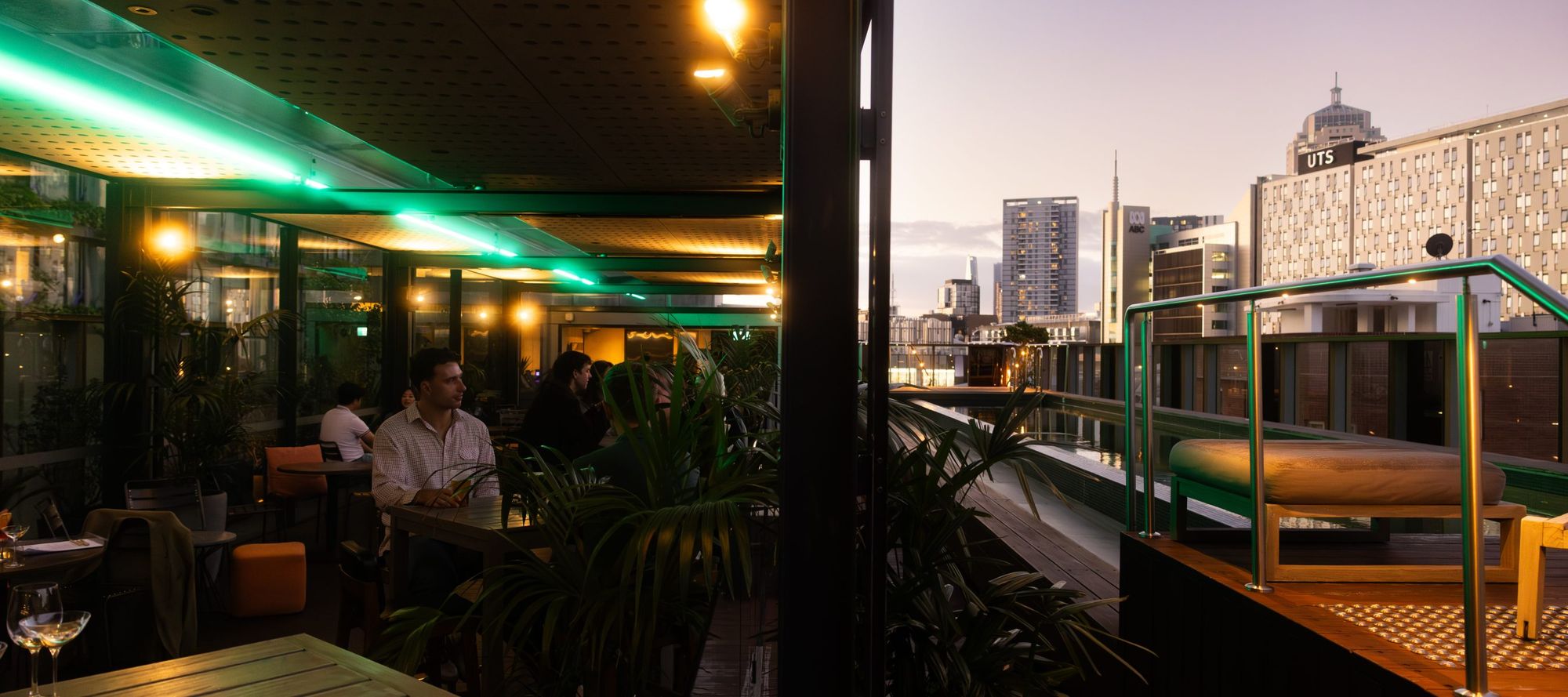 Rooftop bar at dusk with people sitting at tables, surrounded by plants and illuminated by neon lights; city skyline visible in the background.