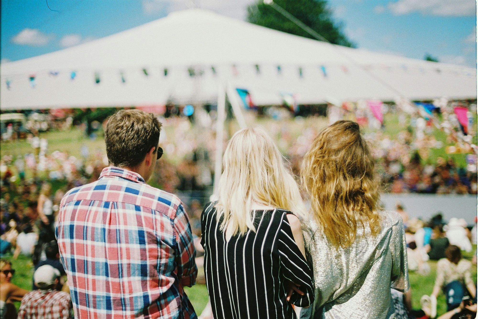 Three people with their backs to the camera at an outdoor festival, facing a large tent and crowd, under a clear blue sky.