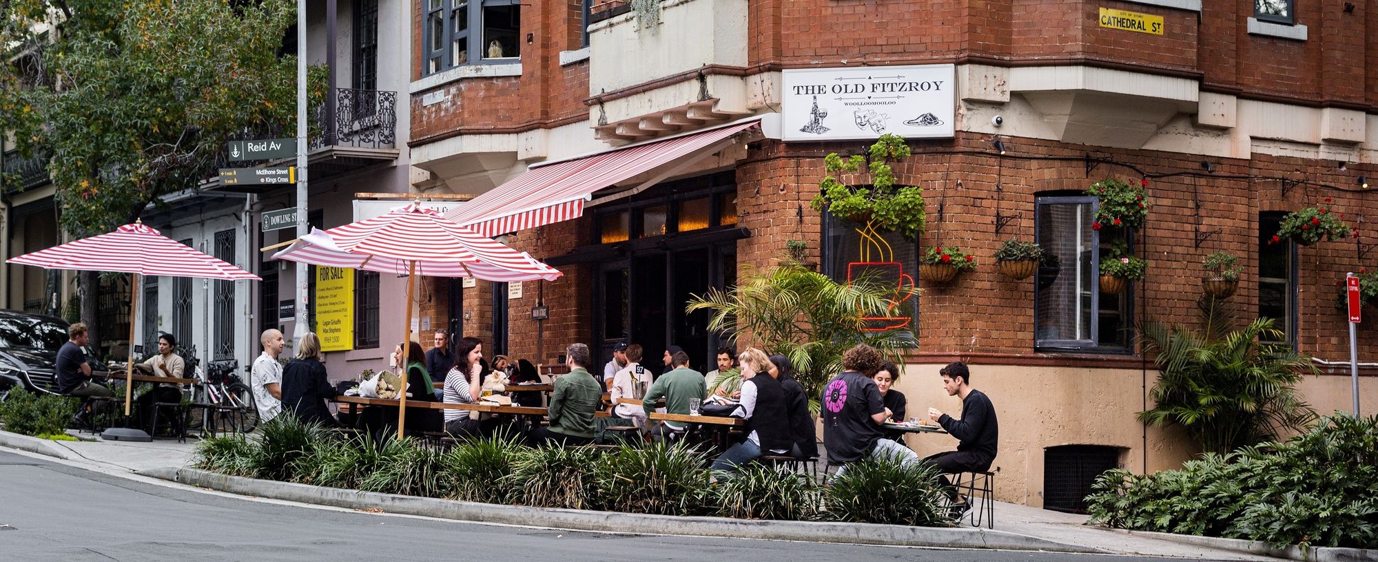 People dining at outdoor tables under red-and-white striped umbrellas outside a brick cafe on a street corner with greenery and potted plants.