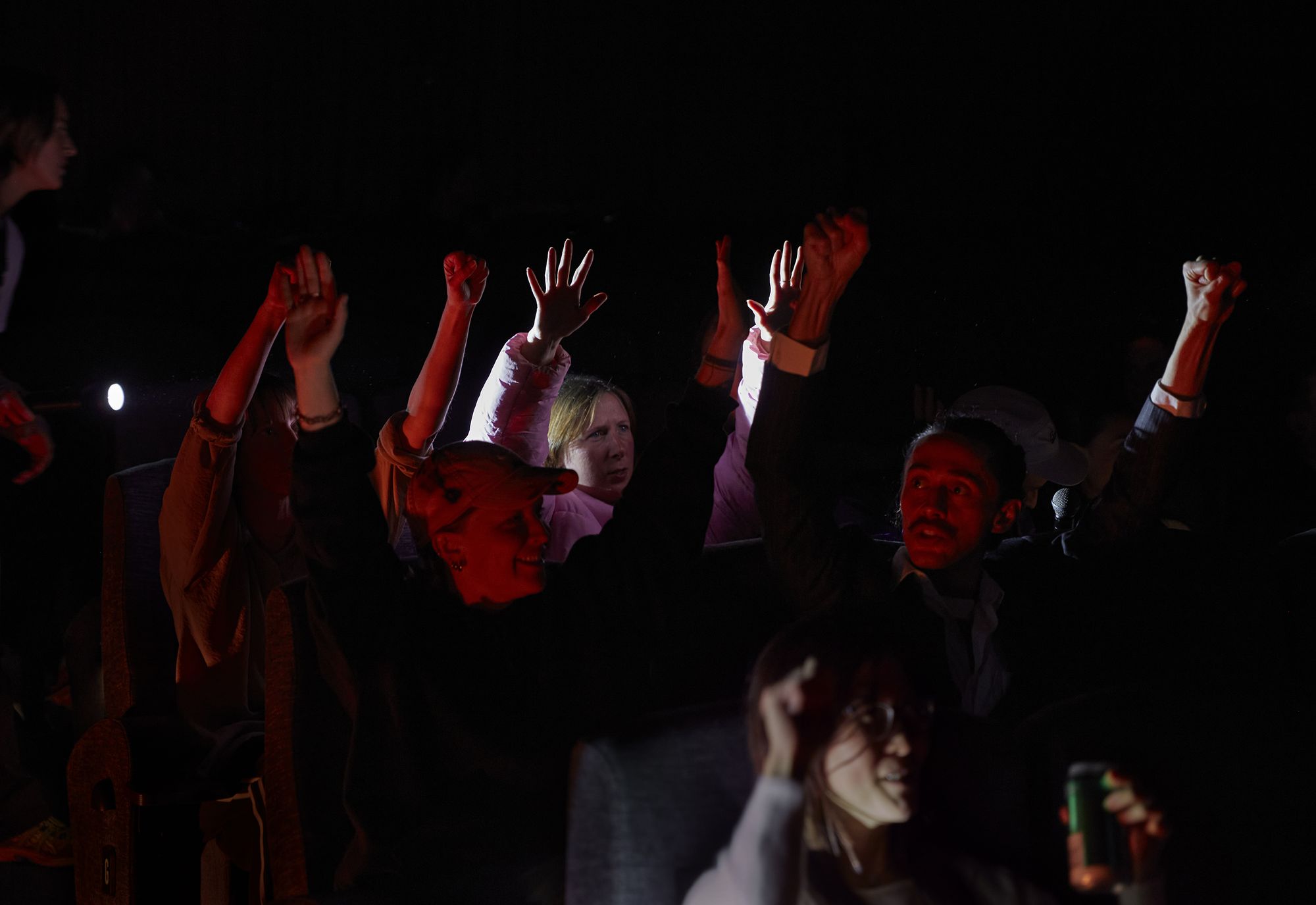 Audience members in a darkened theatre raising their arms, some smiling and illuminated red by theatrical lighting.